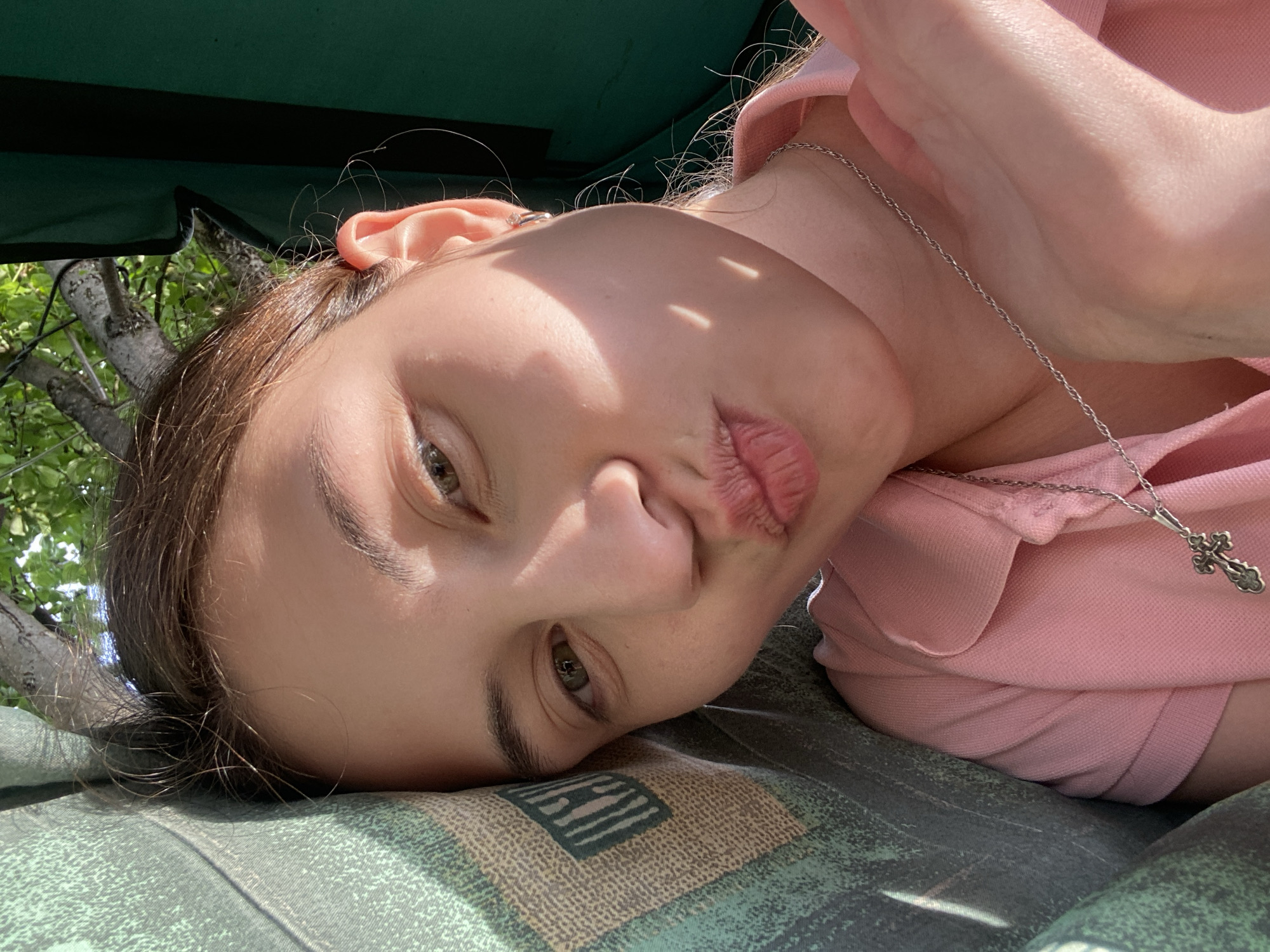 In a close-up, outdoor selfie, a young woman with brown hair pulled back is lying down, her head resting on a textured green cushion. Sunlight streams across her face, casting dappled shadows on her skin and highlighting her green eyes and pink lips pursed in a kiss. She wears a light pink polo shirt and a silver necklace with a cross pendant. The background is out of focus, showing green foliage and a dark green surface, suggesting she is outdoors, possibly in a park or garden. The image appears to be taken in Votkinsk, Russia, on a sunny day.