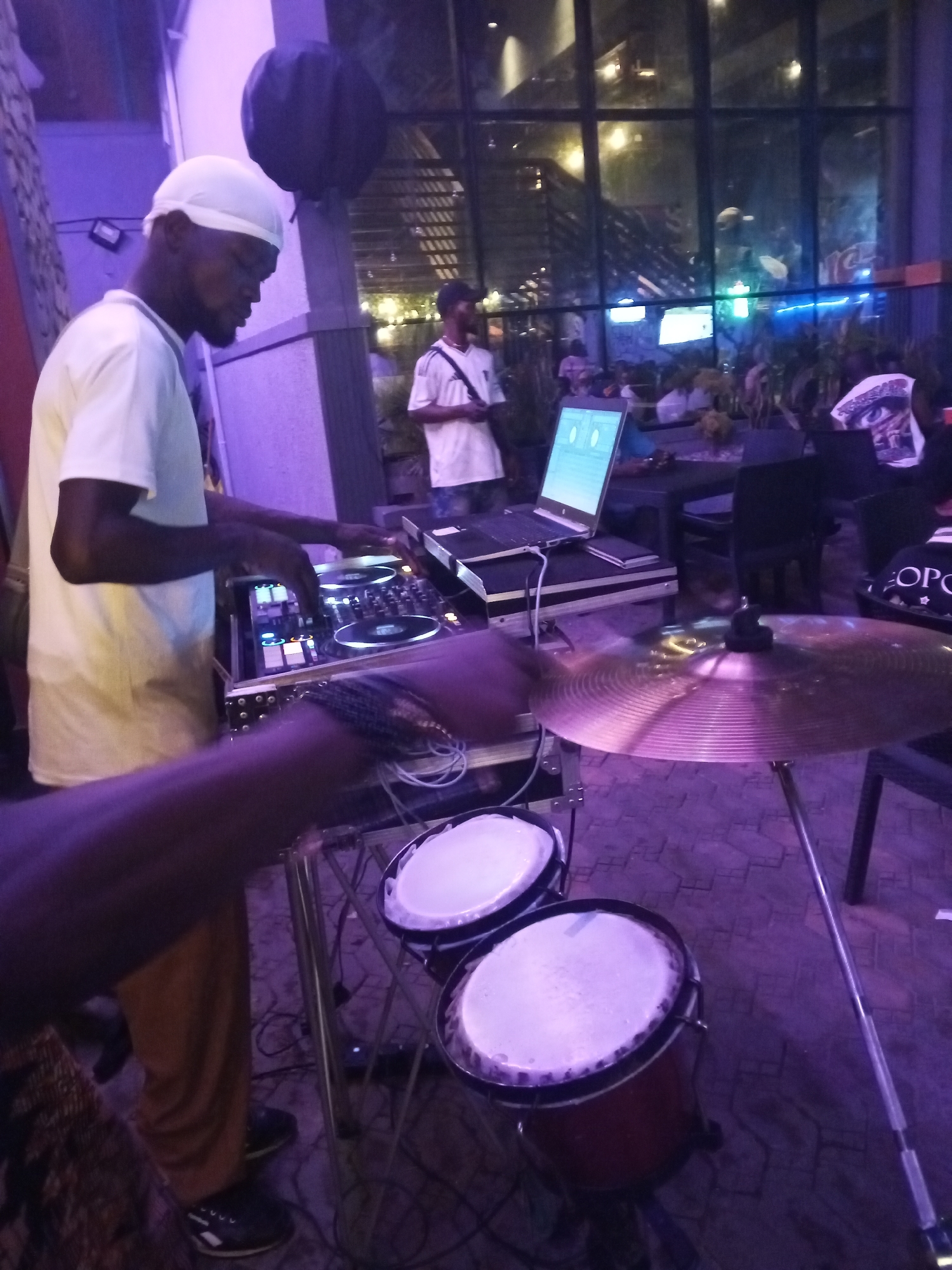 This media file captures a dynamic night scene at an entertainment venue in Lokoja, Nigeria, illuminated by a pervasive purple ambient light. The primary focus is on a Black man, likely a DJ, wearing a white headwrap and white t-shirt, engrossed in mixing music. His hands are positioned over a professional DJ controller, which features illuminated buttons and jog wheels. An open laptop, displaying an interface, rests on a stand beside the controller.
In the foreground, a person's arm, adorned with a bracelet, is visible near a gleaming crash cymbal, suggesting the presence of a live percussionist. Two small, reddish drums with white heads, possibly bongos or small congas, are placed on the tiled floor below the cymbal stand, adding to the musical setup.
Another man in a white t-shirt and dark cap stands further back, observing the activity. Through large windows in the background, reflections and distant lights indicate it is dark outside, and blurred figures suggest other patrons or staff within the venue. Tables and chairs are visible, confirming a lounge or club setting. One individual in the background wears a t-shirt with "OPO" and a stylized eye or face graphic partially visible. The overall scene depicts an energetic musical performance, combining a DJ set with live percussion, in a vibrant, nighttime atmosphere.