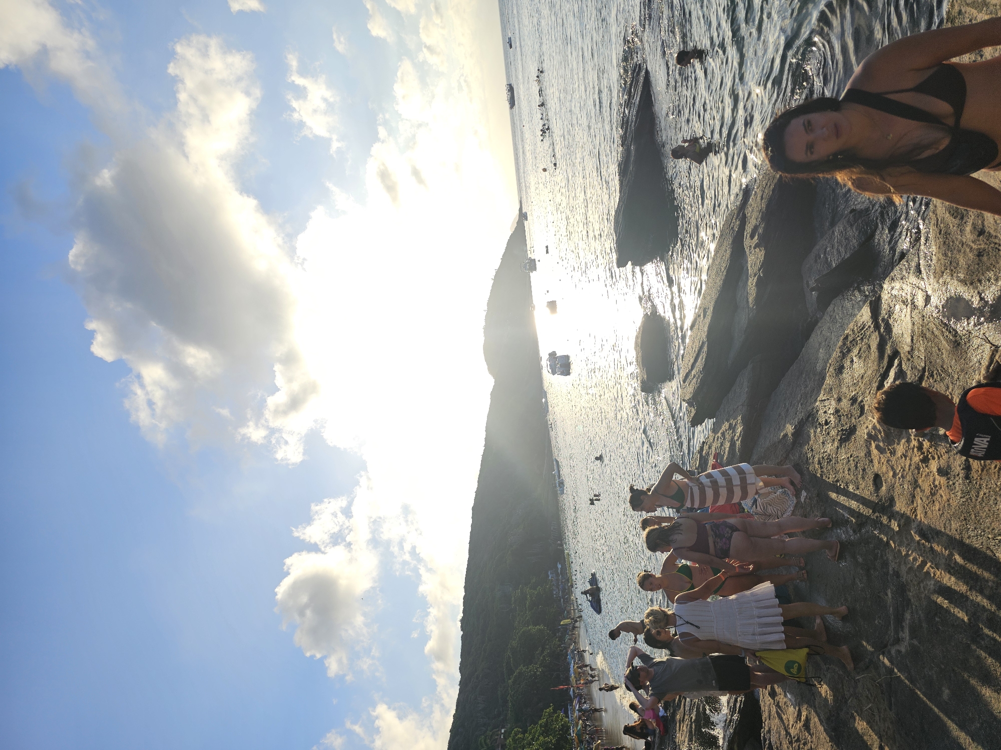 A slightly tilted image captures a sunny beach scene in Armação dos Búzios, Brazil. In the upper right corner, a woman in a dark bikini looks towards the camera from atop rocky terrain. To her left, the ocean stretches out, dotted with swimmers and boats. A group of people stand near the water's edge, facing towards the sea. The shoreline curves into the distance, meeting a tree-covered hillside that rises towards the middle of the frame. The sky, in the upper left, is a mix of blue and white, with clouds scattered across the horizon. The sun's glare shines through, casting a bright glow across the water and sky.