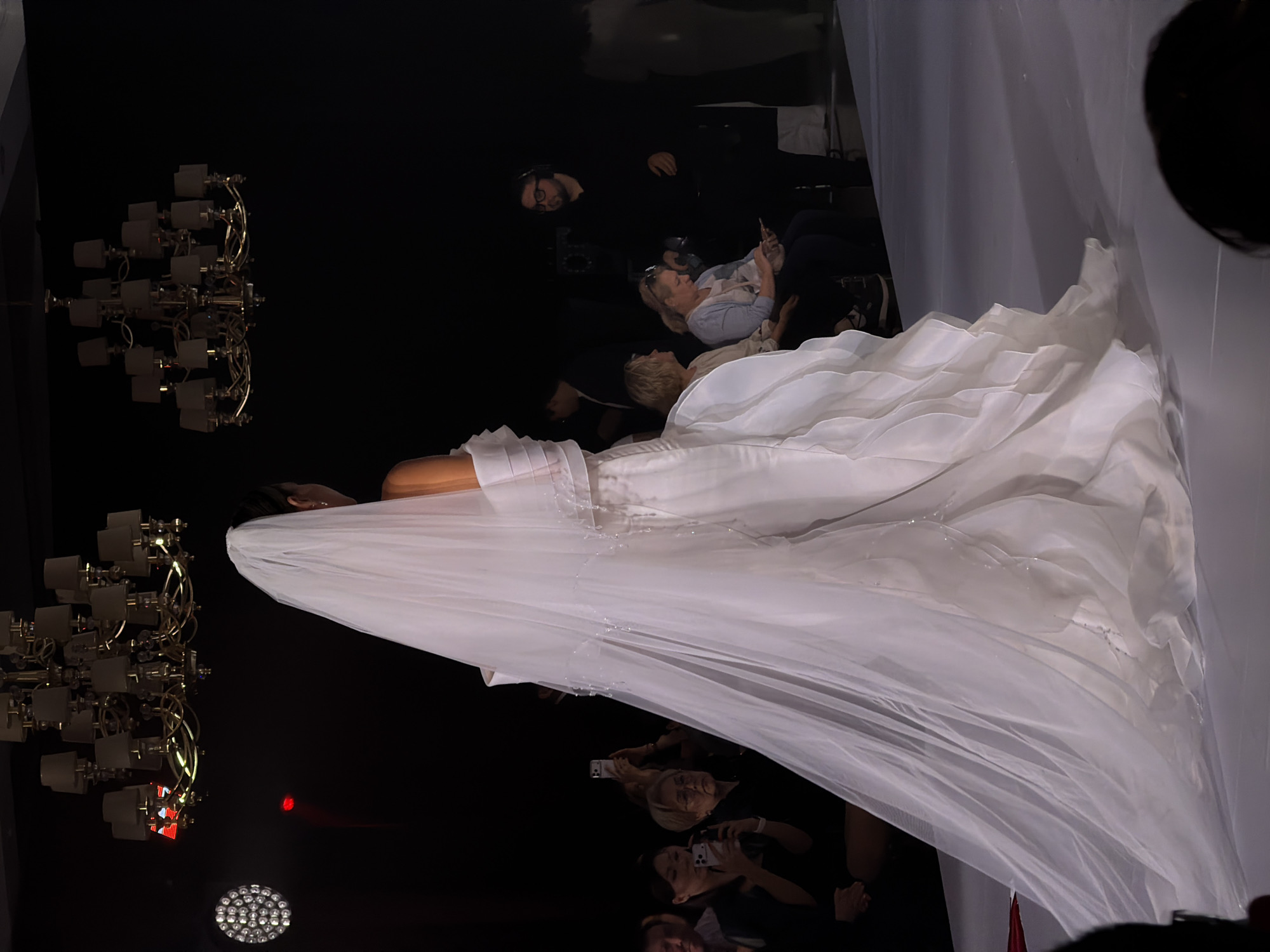 A model in a white gown is posing on a runway, while a crowd of people is taking photos of her. There are chandeliers and stage lights in the background. The location is a fashion show, possibly in a city, but the specific city and location are unknown.