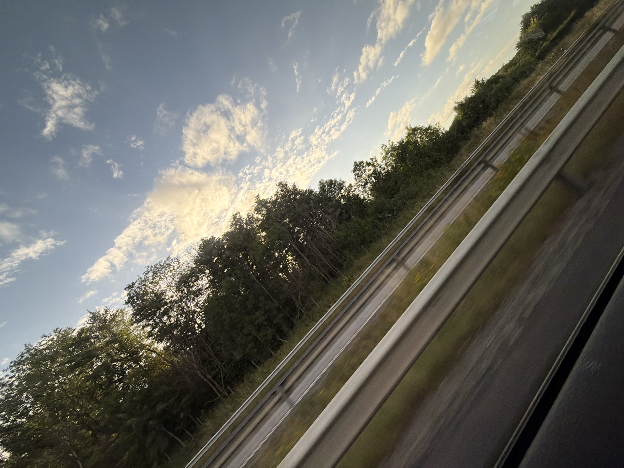 The image shows a view from a moving vehicle, likely on a highway, looking out towards a tree-lined embankment. The perspective is from a low angle, suggesting the camera is near ground level. The road has a metal guardrail in the foreground, which is blurred due to motion. Beyond the guardrail, the embankment is covered with dense green trees and vegetation. The sky above is blue with scattered white clouds, some of which are illuminated by the setting or rising sun, giving them a warm, golden hue. The image is tilted, adding a sense of movement. There are no people or specific landmarks visible that would definitively place it in Kungsbacka, Sweden. The scene is a generic highway landscape.