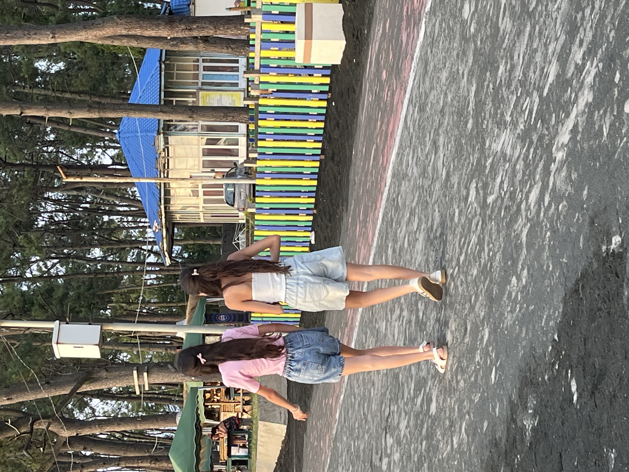 Two young females, viewed from the back, are walking away from the camera on a paved path. The female on the left wears a pink t-shirt and blue denim shorts, with white sandals. The female on the right wears a white strapless top and light blue denim shorts, with tan and white sneakers. They are walking past a colorful fence made of vertical green, yellow, and blue planks. Behind the fence is a building with a blue roof and glass windows. Trees with tall, thin trunks are visible behind the building and fence. In the background, to the left of the frame, are green tents and people sitting at tables. The overall environment appears to be a coastal area, likely in Ureki, Georgia, a resort town known for its magnetic black sand beaches.