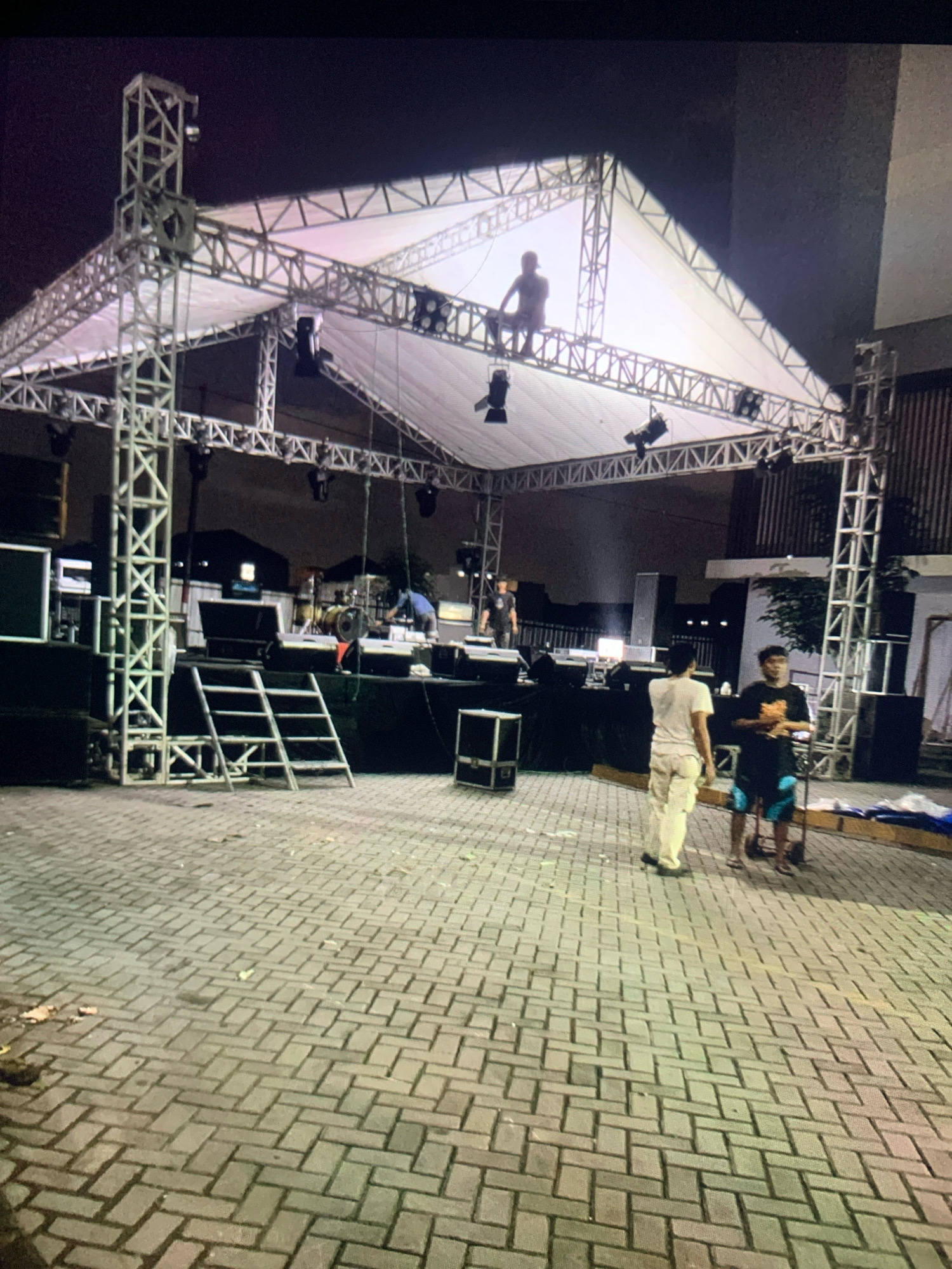 The image shows a temporary outdoor stage setup at night in what appears to be the city of Tawing, Indonesia. The stage is constructed from metal trussing and covered with a white fabric roof. Several stage lights are attached to the trussing. Below the stage, there are large speakers and other audio equipment. Two individuals are visible on the stage, one sitting on the trussing and another standing. In front of the stage, two more individuals are standing on a paved area. The ground is covered with interlocking brick pavers, and debris is scattered on the ground. A ladder leans against the stage structure. In the background, there are buildings and fences. The overall scene suggests preparation for an event, likely a concert or performance.