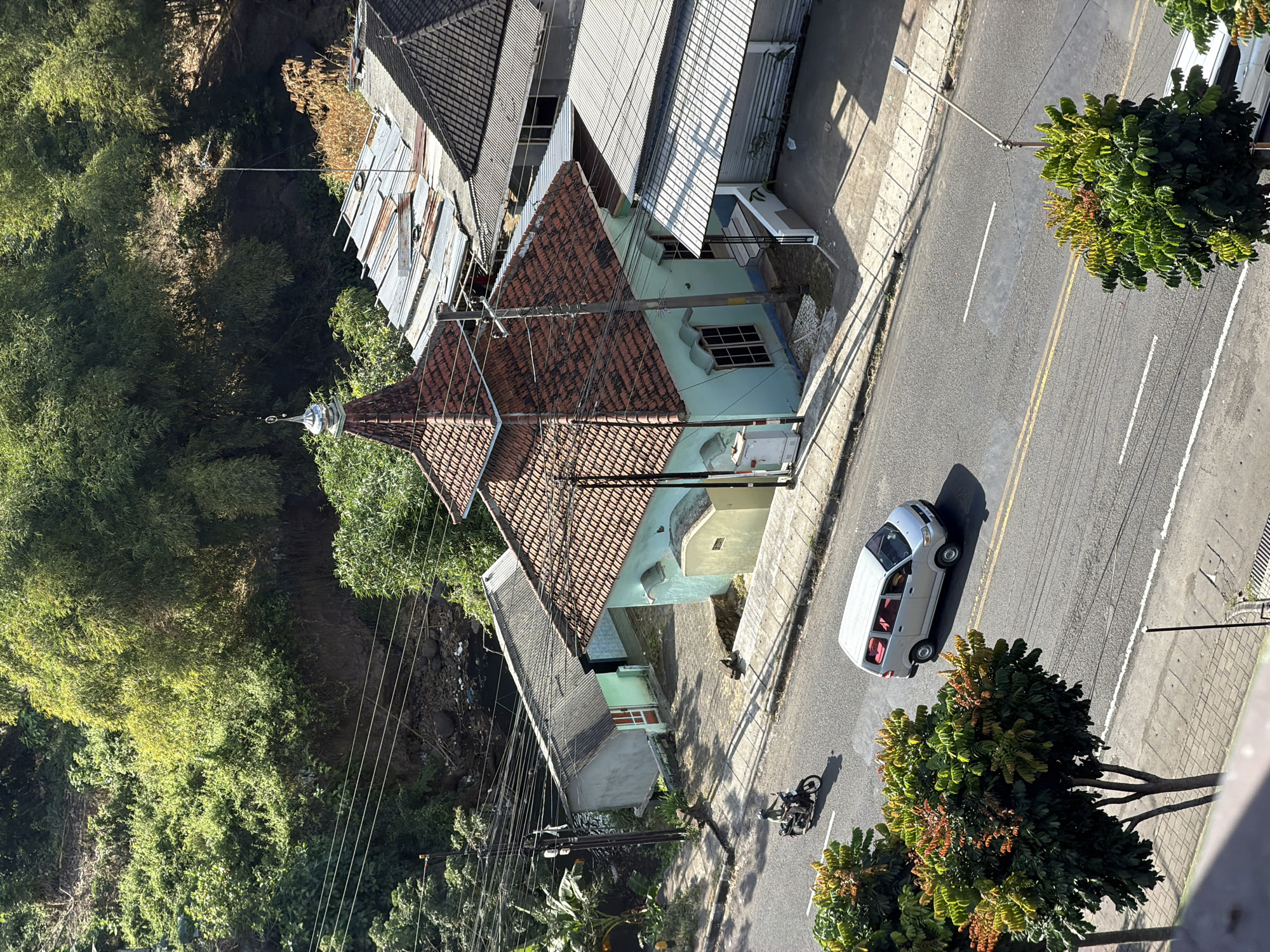 An elevated, eye-level, static, medium shot captures a scene in the city of Dadapan, Indonesia, on a sunny day. The focal point is a light blue building with a terracotta-tiled, conical roof, topped with a silver finial. This structure, likely a mosque, is situated on a slight incline behind a paved road. A white minivan is driving on the road, and a motorcycle with a rider is also present. Trees with lush green foliage, some with reddish-brown accents, line the sides of the road and surround the buildings, creating a vibrant natural setting against the urban landscape. The background features a densely vegetated hillside, indicating the city's proximity to nature. Utility poles and overhead wires are visible, common elements in Indonesian streetscapes. The overall impression is a snapshot of daily life in a tropical urban environment.