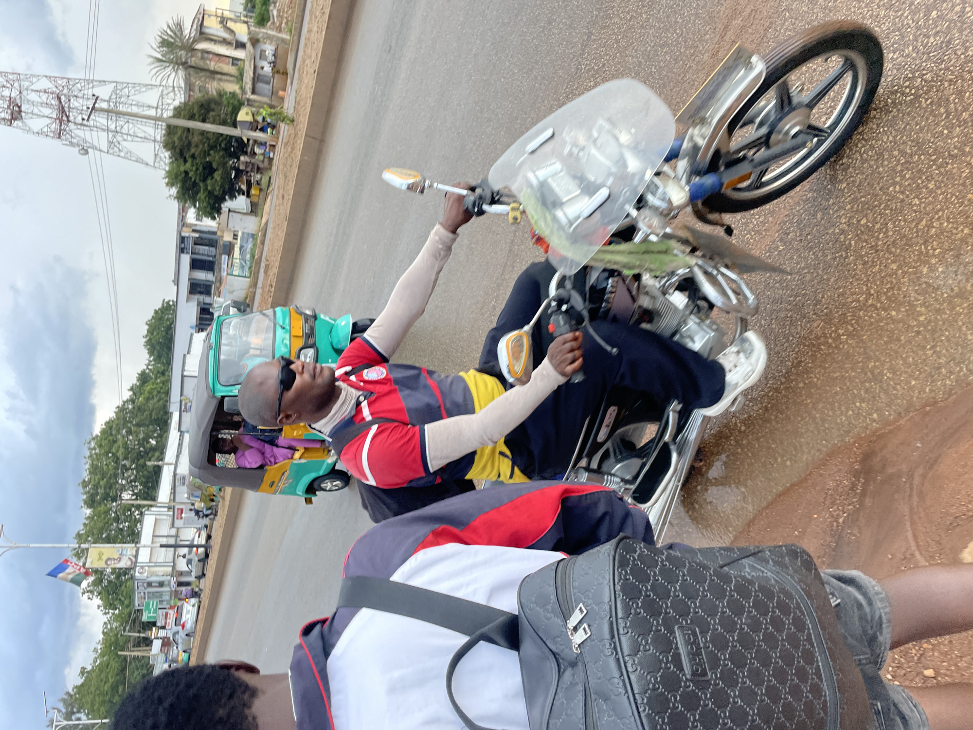 A medium shot captures a man riding a motorcycle on a wet, reddish-brown asphalt road, likely in Jos, Nigeria. The man, wearing sunglasses, a red, yellow, and black striped polo shirt with light beige sleeves, and dark pants, is positioned in the center-right of the frame. His left hand is on the handlebars, and his right arm is extended slightly. A clear windshield is attached to the front of the motorcycle. The motorcycle's front wheel is turned slightly to the left, and the spokes are visible. A green and white cloth is draped over the front of the bike. In the background, a turquoise and yellow auto rickshaw with a woman wearing a purple garment is driving away. Further back, several cars, buildings, trees, and a large telecommunications tower are visible under a cloudy sky. On the left side of the frame, the back of another person wearing a black backpack with the Gucci logo is visible, along with part of a blue and white jacket and grey shorts. The road surface appears wet and reflective, with some gravel visible.