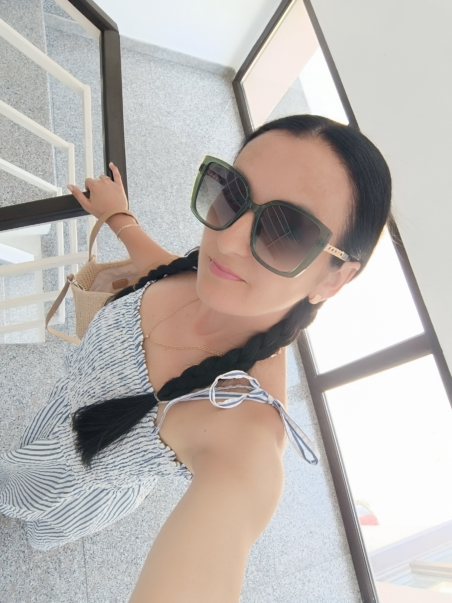 A woman with dark hair in two braids is taking a selfie. She is wearing large green sunglasses, a blue and white striped dress, a gold necklace, and a woven purse. She is standing on a speckled floor next to a white railing of a staircase. A large window with a reflection is to her right. The woman is wearing a ring on her left hand.