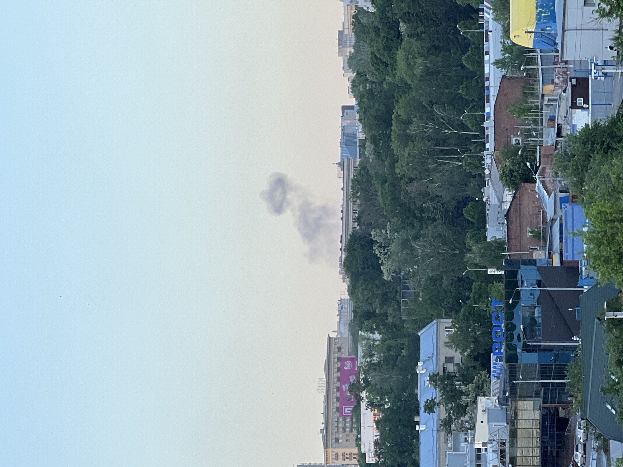 The image depicts a skyline in Kharkiv, Ukraine. In the foreground, there are residential buildings, some painted in blue and white. A dense line of trees stands between the buildings and the more distant cityscape. Further back, there are taller buildings and skyscrapers. Smoke is visible in the sky. The sky itself is a pale blue-grey.