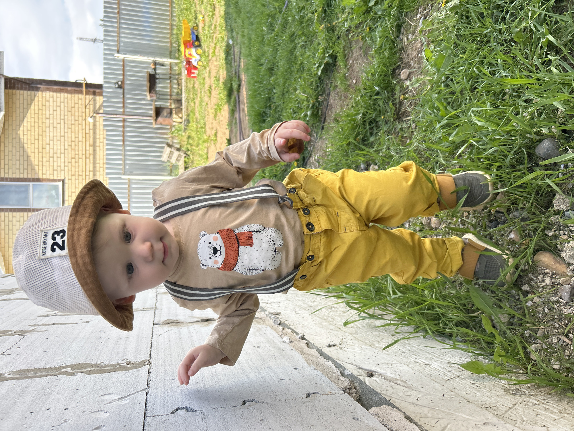 A toddler wearing a light brown shirt with a polar bear graphic, yellow pants with suspenders, and a brown-brimmed hat labeled "KOBE BRYANT 23" is lying on a concrete surface. The background includes a brick building and a corrugated metal fence. Grass and a few small rocks are visible. The exact location within Chaikovsky, Russia is impossible to determine with this image alone.