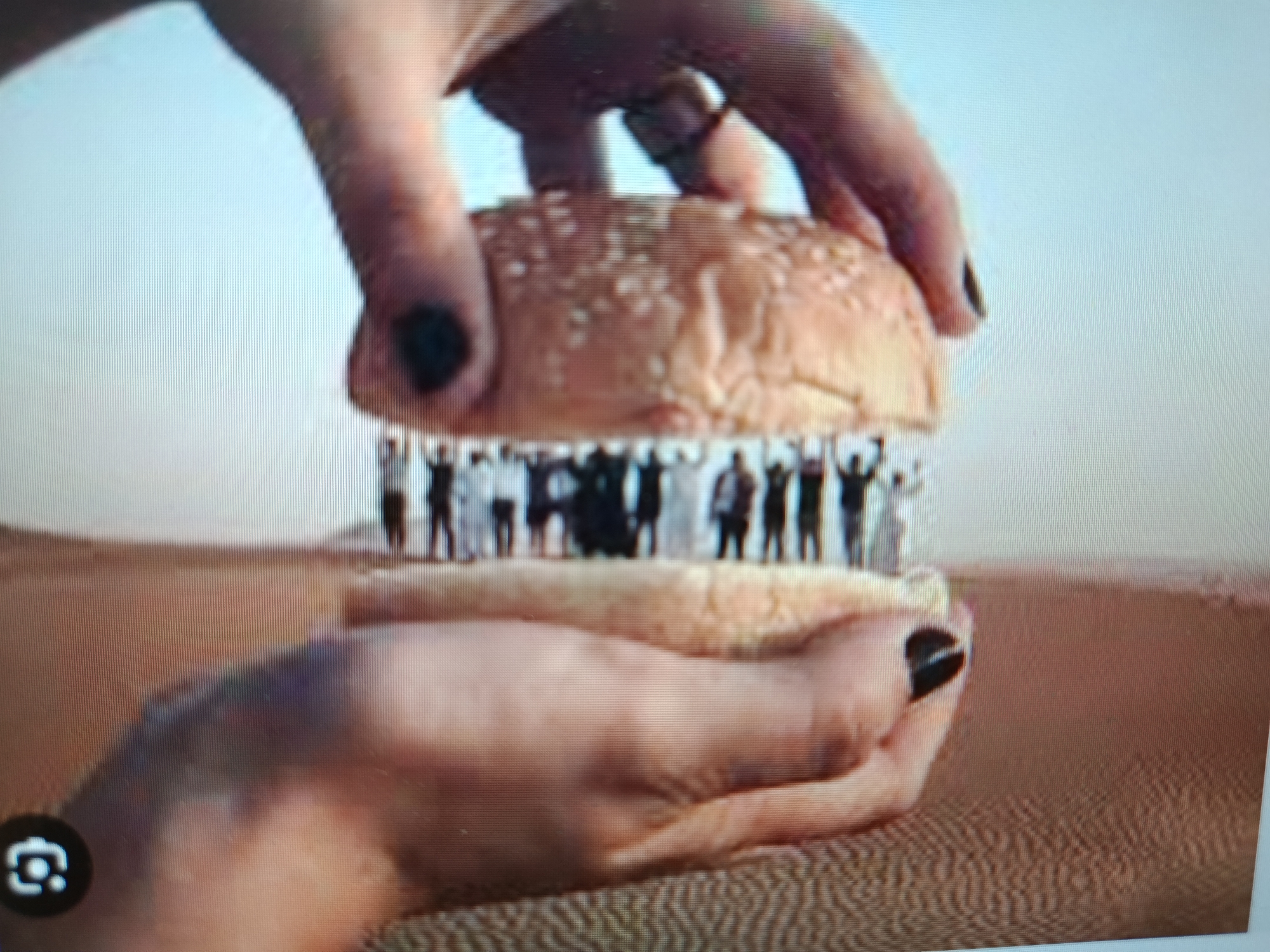 This image shows a close-up of a hamburger bun being held by two hands. Inside the bun, a miniature scene is depicted with a group of small figures standing with their arms raised. The background appears to be a desert landscape. The overall impression is a surreal or artistic composition. There is no discernible information within the image that would associate it with the city of Tashkent, Uzbekistan.