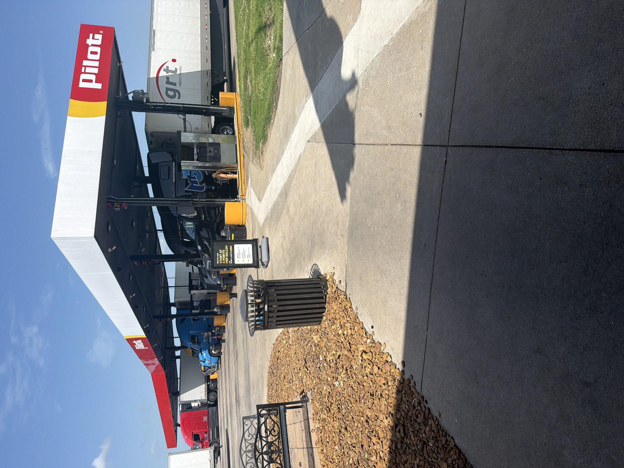This is an eye-level, outdoor, medium shot of a Pilot gas station with multiple semi-trucks refueling under a covered canopy. The sky is clear and blue with a few wispy clouds. The Pilot logo, a red rectangle with a yellow arc and white text, is prominently displayed on the white canopy. Several fuel pumps are visible, with one black semi-truck hooked up and another blue semi-truck parked nearby. The ground is paved concrete with some areas of gravel and grass. A black metal trash can and a decorative metal bench are positioned on the gravel area in the foreground. The overall scene depicts a typical truck stop environment on a sunny day in Hayti, United States of America.