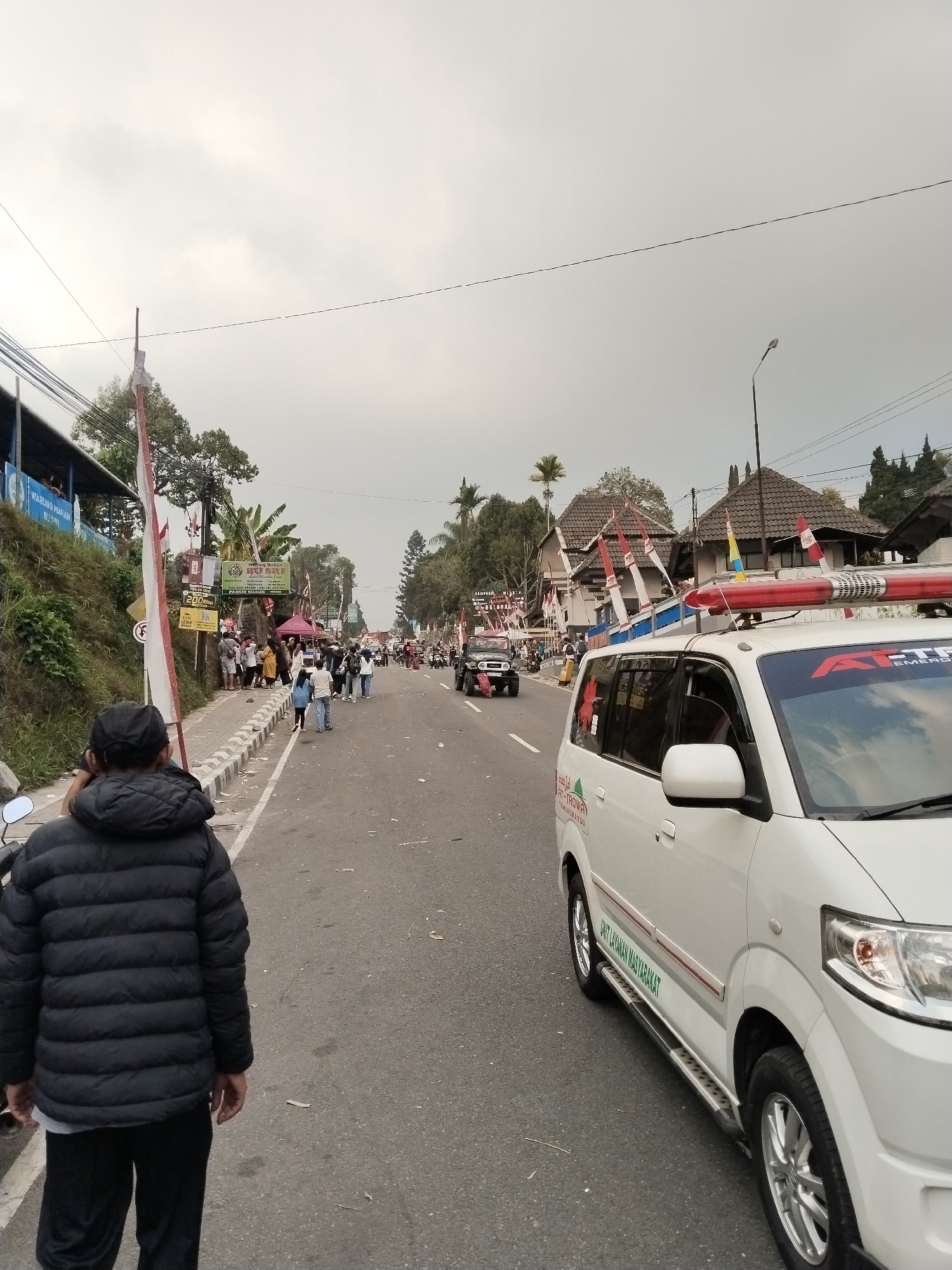 In Tawangmangu, Indonesia, a street scene unfolds under an overcast sky, likely during an event or gathering. In the foreground, a person in a black puffer jacket and black pants stands facing away from the camera, appearing to be either walking or waiting. To the right of the street, a white vehicle, possibly an ambulance or a service van, is parked, equipped with a flashing red light bar on its roof and text in Indonesian on its side. The vehicle's windshield displays a red decal with "ATI-TAWA" and "EMERGENCY".
The street itself is paved and lined with a sidewalk. Along the left side of the road, a grassy embankment rises, topped with buildings and trees. Banners and Indonesian flags are visible, attached to poles and buildings, suggesting a festive or official occasion. Numerous people are present, walking on the sidewalk and along the edge of the street, some in groups, observing the surroundings.
Further down the road, other vehicles are visible, including a dark-colored jeep-like vehicle and more parked cars. The background reveals more buildings with tiled roofs and lush green trees, characteristic of a hilly or mountainous region. The overall atmosphere suggests a community event or a procession, with a mix of public participation and official vehicles present. The presence of a warung (small shop) with a blue awning on the left side indicates commercial activity along the route.