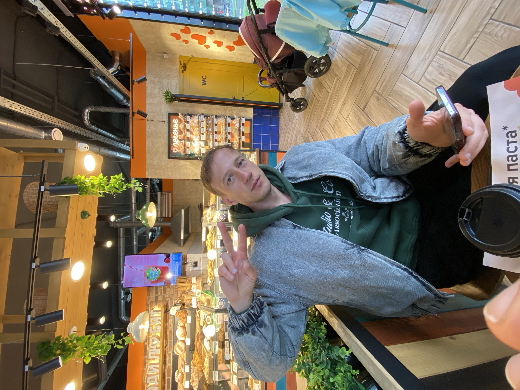 A man is posing for a photo in a café in Murino, Russia. He's wearing a gray denim jacket over a green hoodie. He is holding a smartphone in his right hand and making a peace sign with his left hand. A disposable coffee cup is visible to his right. The café has wooden floors, a yellow restroom door, and displays of baked goods. A stroller sits nearby.