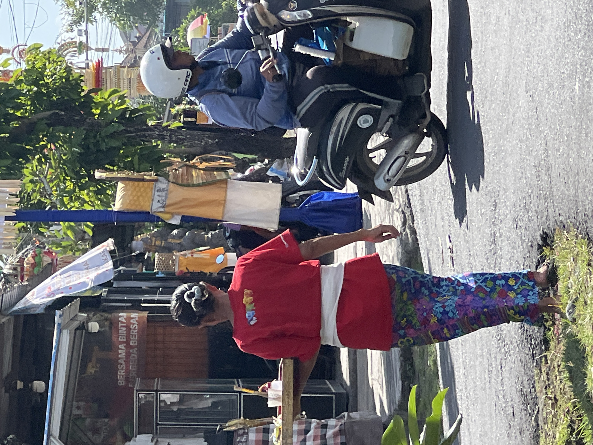 In North Kuta, Indonesia, on a sunlit day, a woman in a red t-shirt and a vibrant floral sarong walks on the side of a paved road, carrying a wooden tray with offerings. A man on a black scooter, wearing a blue jacket and a white helmet, rides past her on the road. Behind them, a building with signage and decorative banners suggest a commercial area. Green foliage and trees add to the tropical atmosphere.