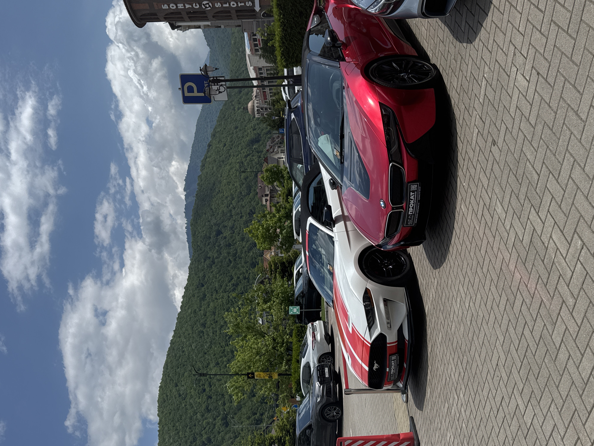 The image captures a row of parked cars on a paved surface, likely a parking lot, with a backdrop of lush green mountains under a partly cloudy sky. The orientation of the image is vertical, suggesting it might have been taken with a phone held upright.
In the foreground, the most prominent subjects are several cars. Closest to the viewer and on the right side of the frame is a red and white sports car with racing stripes. Further down the row, a sleek red sports car is visible. Other cars, including a dark-colored SUV and a white SUV, are also parked in the line. A blue parking sign with a white "P" is mounted on a pole.
The scene appears to be set in a scenic location, possibly a resort area, given the presence of the mountains and what looks like hotel buildings in the background. The weather seems pleasant, with a bright sky and scattered clouds, suggesting it is daytime. The overall atmosphere is calm and still, with no people visible.
Text is present on the license plates of some cars and on a sticker on the front of the red and white sports car. The sticker displays the word "ПРОКАТ" in Cyrillic script, which translates to "RENTAL," indicating that the car is likely available for rent. There is also a website address and a phone number on the sticker. Above the cars, text is visible on the facade of a building, appearing to be in Cyrillic script as well.