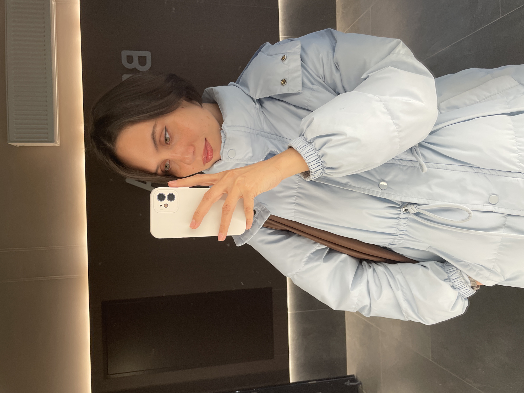 A woman is taking a selfie in what appears to be a retail store or boutique. She is wearing a light blue, puffy winter coat with a drawstring waist. She is holding a white smartphone in her right hand, taking the picture. The background includes a dark, smooth surface, likely a wall, with the partial letters "BA" visible. To the left, there is a vertical, illuminated design element. The floor is visible and seems to be tiled in a dark gray color. The location cannot be determined to be Izhevsk, Russia based on this image.