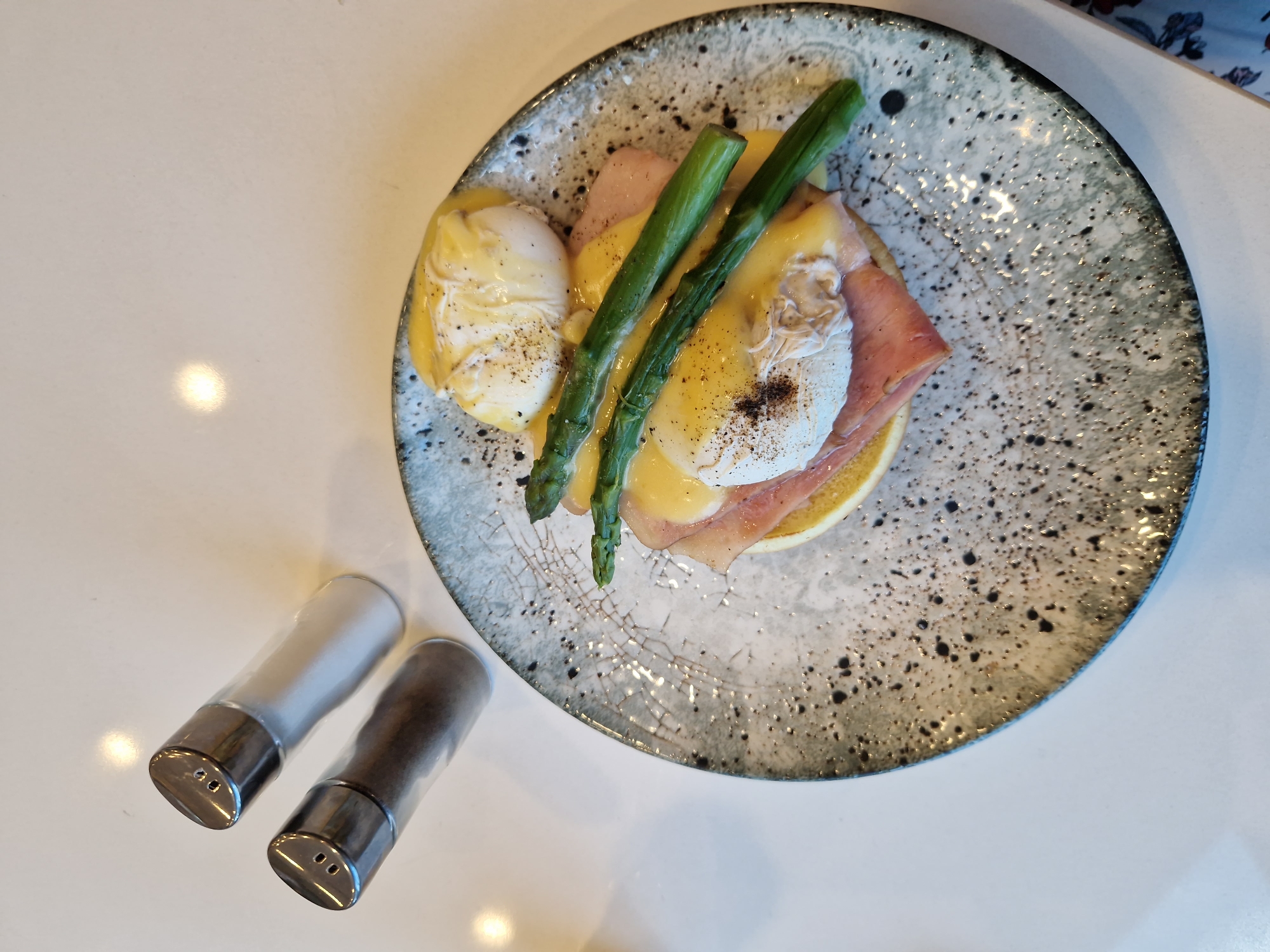 A close-up, overhead shot shows a plate of Eggs Benedict on a speckled ceramic plate, positioned on a white countertop. The dish features two perfectly poached eggs, generously coated in hollandaise sauce, and topped with a sprinkling of black pepper. Beneath the eggs are slices of ham and a thick piece of toasted bread. Two spears of bright green asparagus are artfully placed on top of the hollandaise. To the upper left of the plate are two glass salt and pepper shakers with silver lids. The lighting creates soft shadows and highlights, indicating an indoor setting, likely a restaurant or cafe. The overall scene is clean and well-lit, emphasizing the appetizing presentation of the food. The image does not depict any people or specific architectural details that would indicate a precise location in Sofia, Bulgaria.