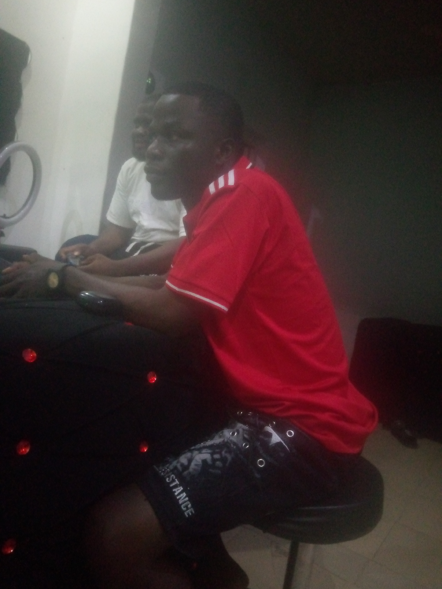 An interior scene depicts two dark-skinned males seated in a dimly lit room. In the foreground, a male subject is seated facing left, wearing a red short-sleeved polo shirt with white stripes on the collar and sleeves, and dark shorts with a "STANCE" graphic and silver eyelets. A dark watch with a gold-colored bezel is visible on his left wrist. His left hand rests on a dark, cushioned surface embellished with several small, red, circular illuminated elements. He is seated on a dark, circular stool.
Behind him, to the left, a second male subject is partially visible, wearing a white short-sleeved t-shirt. His hands are clasped in his lap, possibly holding a small object.
The background features a light-colored wall on the left and a dark-colored wall on the right. A circular ring-like object, possibly a light or stand, is visible against the light wall. The floor in the bottom right corner is light-colored and tiled. A dark object, possibly a bag, is on the floor against the dark wall. The specific location within the city of Unknown, Unknown, is not discernible from the image.