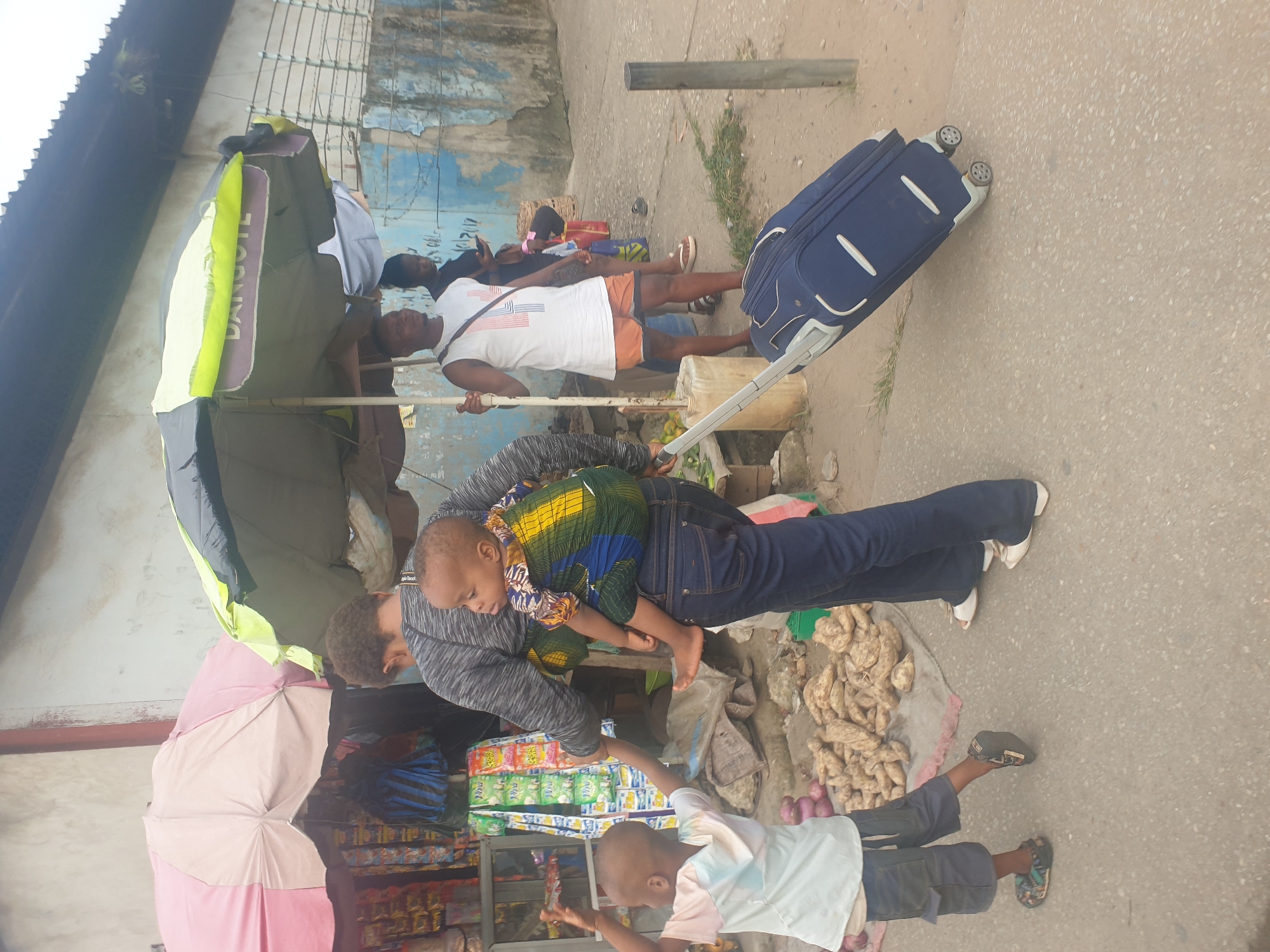 A photograph taken outdoors in Aba, Abia, Nigeria shows several people, a blue rolling suitcase, and roadside commerce.
A woman, wearing a gray long-sleeved shirt and blue jeans, bends over and appears to be handing something to a young boy who is reaching up towards her. The woman carries a baby on her back. A pile of what appears to be potatoes and onions is next to her. To the left of the woman is a shop with pink, white, and dark blue tarps or umbrellas. A person stands underneath it.
Further back and to the right, a man wearing a white tank top with red and blue stripes, and orange shorts, stands near a man with a black tank top. They both hold onto a long pole, supporting an umbrella. A blue rolling suitcase is nearby. The background features a dilapidated building.
