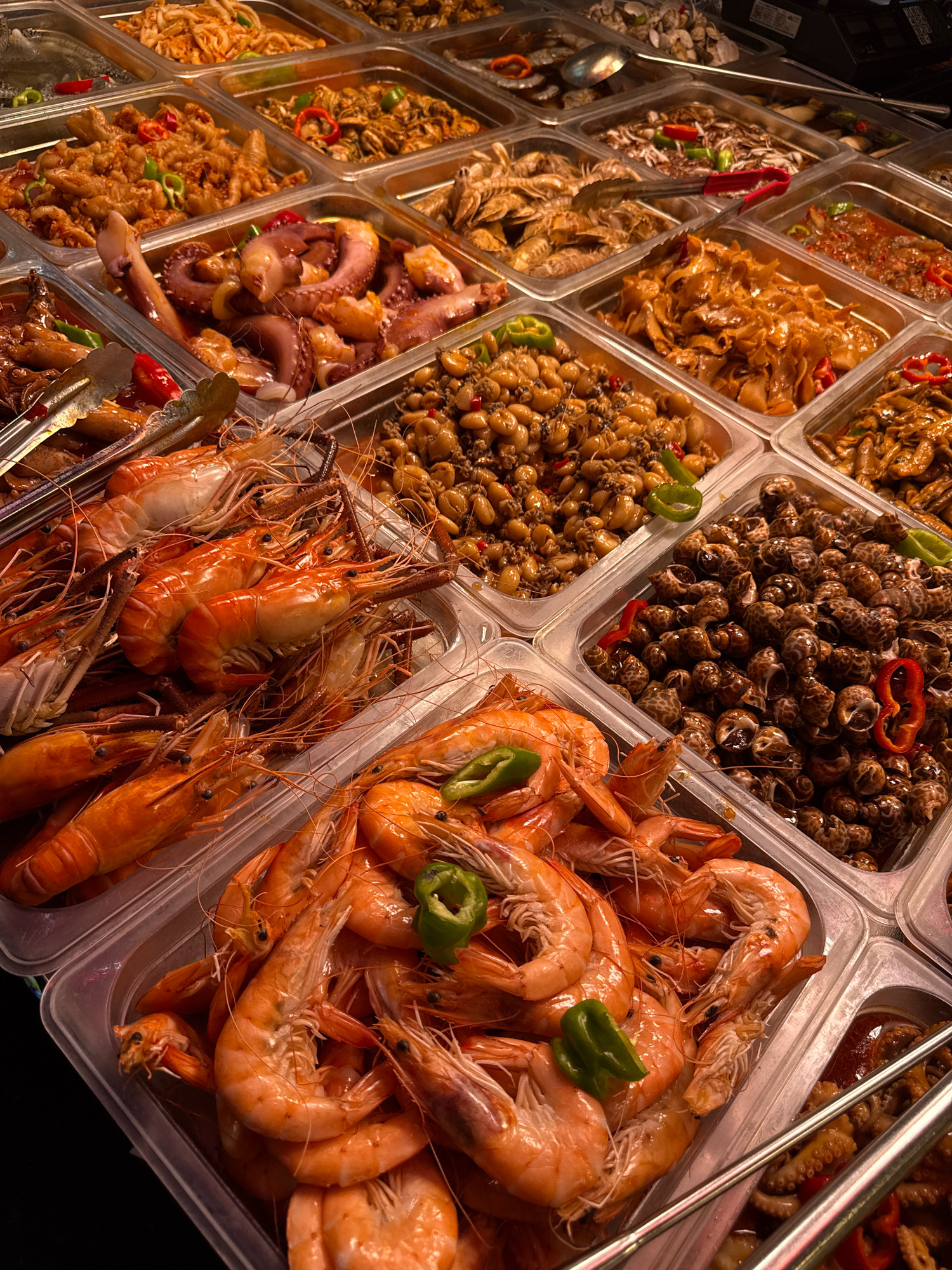 The image depicts a close-up, overhead view of a food stall display in Yiwu, China, likely in a market. The stall is filled with a variety of seafood dishes presented in rectangular, clear plastic trays. Prominently featured are large, cooked shrimp with their shells on, arranged in several trays in the foreground and middle ground. Other dishes include cooked octopus tentacles, small snails, and what appear to be various stir-fried or braised seafood items with vegetables and chili peppers. Tongs are visible in some of the trays, suggesting a self-serve or buffet-style setup. The lighting appears to be artificial, casting a warm glow on the food. The overall impression is one of a diverse and abundant seafood offering at a food vendor.