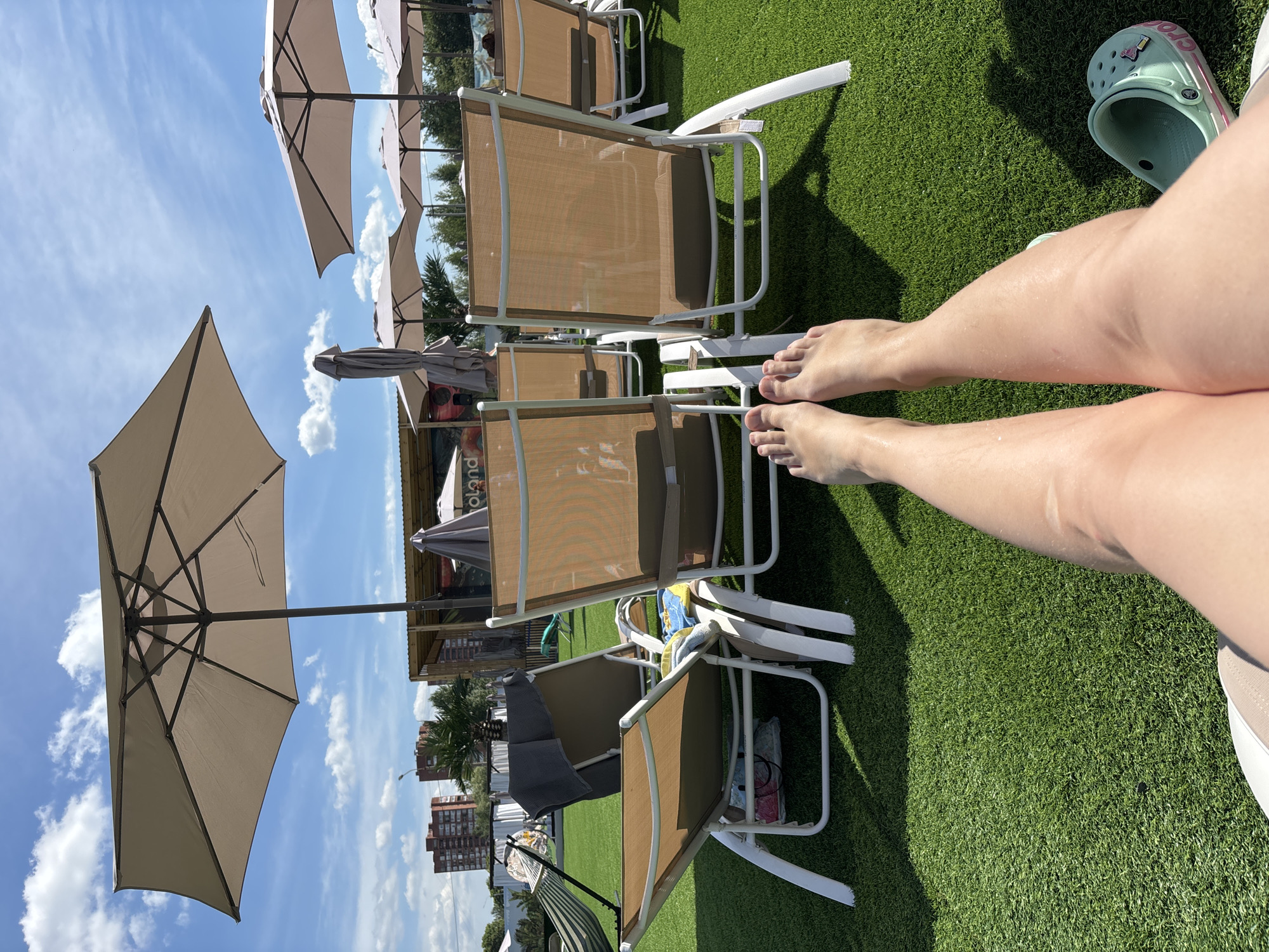 A first-person perspective image shows a person's legs and feet relaxing on a grassy area, possibly a park or resort, with lounge chairs and umbrellas in the background under a blue sky with white clouds. The person is wearing Crocs shoes. Buildings and trees are visible in the distance. The location is identified as Yaroslavl, Russia.