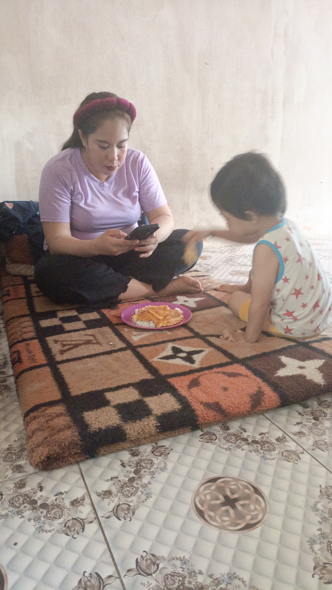 A woman in a purple t-shirt and dark pants, wearing a pink headband, is sitting on a rug on the floor, looking at her phone. Next to her, a toddler in a white and blue star-patterned shirt and yellow shorts is reaching out towards her. A pink plate with rice and some food on top sits on the rug between them. The scene is indoors, with a plain, light-colored wall behind them and a patterned floor. The environment appears to be a simple living space, possibly in the city of Pejala, Indonesia, given the context.