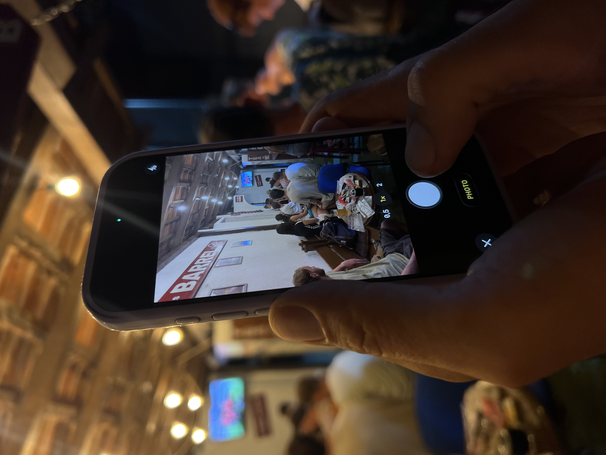 The image shows a person holding a smartphone and taking a photograph of a busy bar. The phone screen displays a scene inside a bar with several people sitting at tables and a sign that reads "BARREL". The background of the phone screen shows a ceiling with exposed wooden beams and light fixtures. The environment outside the phone screen is dimly lit, with some people visible in the background. The phone's camera interface is visible at the bottom of the screen, showing options like zoom levels and a "PHOTO" button. The location is identified as the city of Bar, Montenegro, suggesting this bar is a popular establishment in the area.