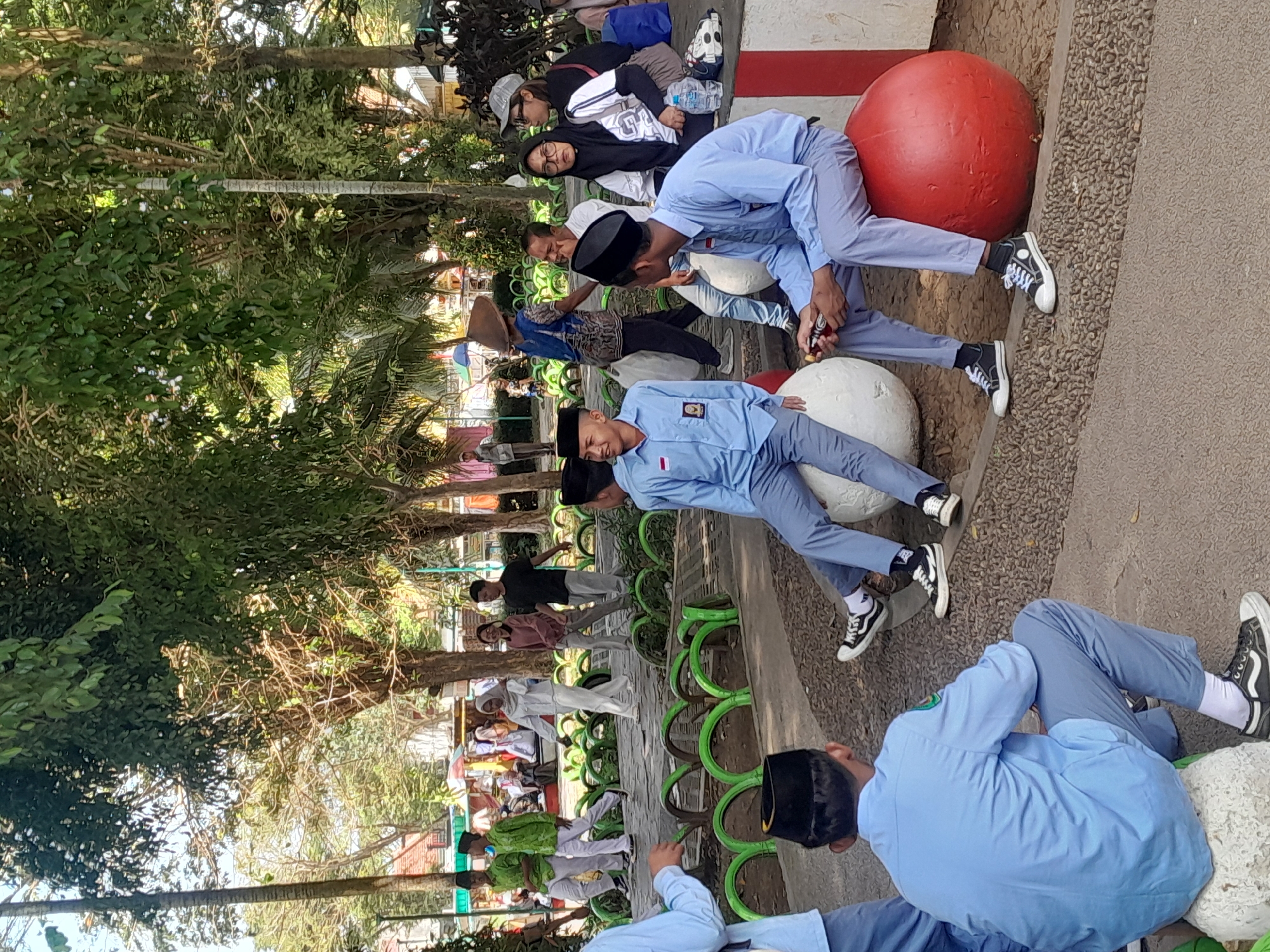 In a public park in Genteng Kulon, Indonesia, several individuals are gathered. In the foreground, two young males in blue uniforms and black peci hats are seated on large, rounded objects, one red and one white. Both are wearing white socks and black Converse-style sneakers. The male on the right is holding a bottle. Behind them, another young male in a similar blue uniform is partially visible, also wearing a black peci and white socks with sneakers.
Further in the background, a group of people are walking. Two young males in green shirts and white pants are walking from left to right. A young female in a white hijab and white clothing walks behind them, followed by a young female in a pink top and grey pants. Another young male in a black shirt and grey pants walks to their right.
Other figures are visible further back, including a person wearing a conical hat and a patterned blue shirt, and individuals sitting on a concrete bench. The environment is characterized by trees with green foliage, a paved pathway, and decorative tire barriers painted green. The lighting suggests it is daytime.