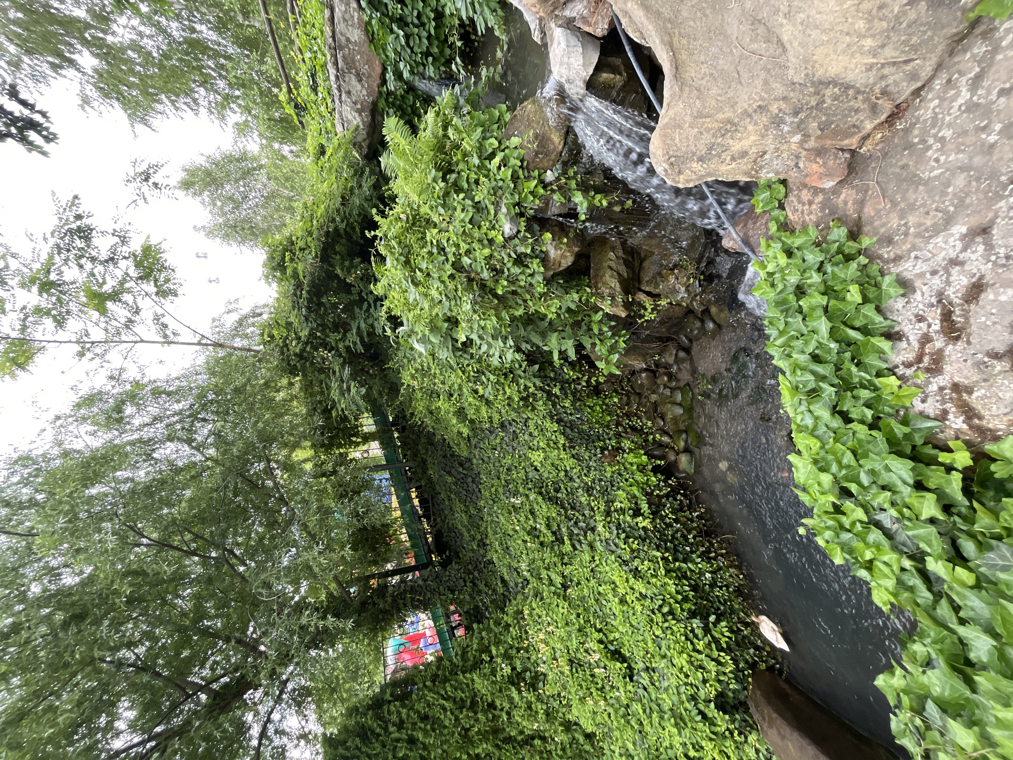 The image depicts a small, artificial waterfall feature constructed with rocks and vegetation. Water cascades over the rocks into a narrow channel, which is partially covered with ivy. The surrounding area is lush with greenery, including trees and dense ground cover. A black metal railing is visible, suggesting a park or landscaped area. In the background, blurred structures and colors indicate the presence of buildings, possibly a playground. The location is Miatli, Russia.