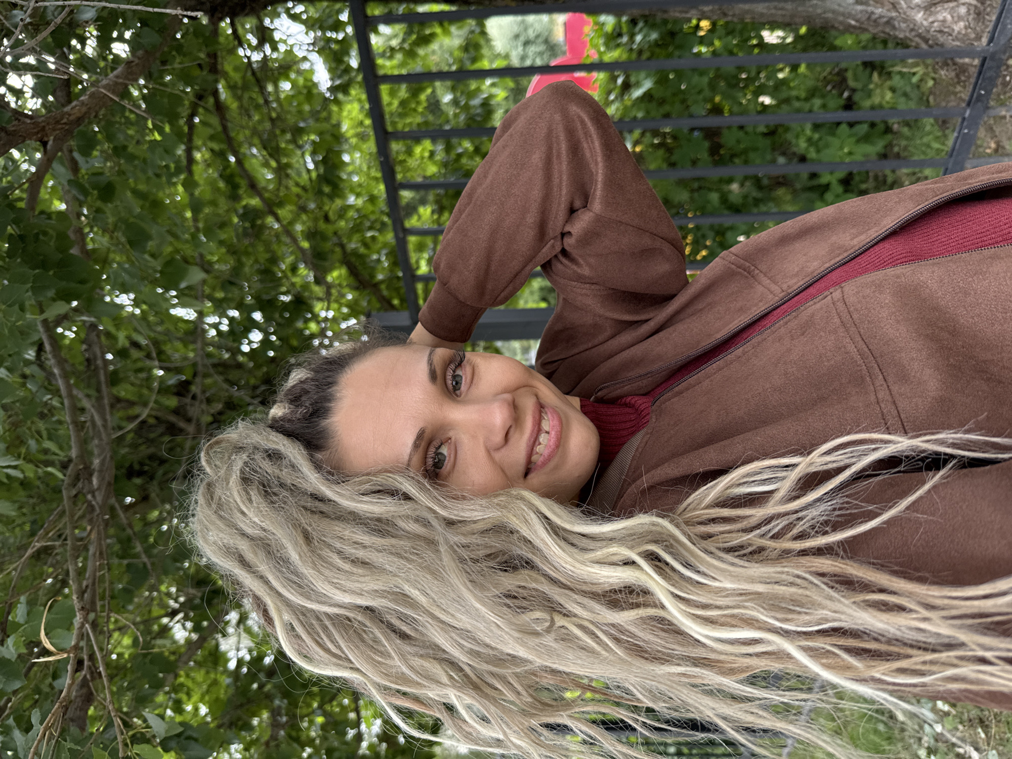 A close-up, waist-up shot features a woman with long, blonde, wavy hair styled in a high ponytail. She is looking at the camera and smiling, with her right hand touching the back of her head. She is wearing a brown jacket over a dark red turtleneck sweater. The background consists of lush green foliage and a black metal fence. The scene appears to be outdoors, possibly in a park or a residential area, with dappled sunlight filtering through the leaves. There are no specific landmarks to indicate the city of Rusanovo, Russia.
