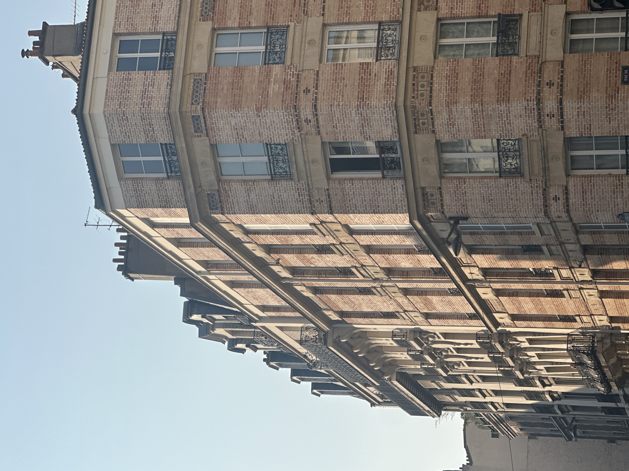 The image captures a close-up of the corner of a Parisian apartment building made of light-colored bricks with a mansard roof. The building has multiple stories with numerous windows, many featuring wrought-iron balconies. The architectural style is typical of Haussmannian buildings found in Paris. The sky is clear and blue, suggesting a sunny day. The building appears to be in a well-maintained condition.