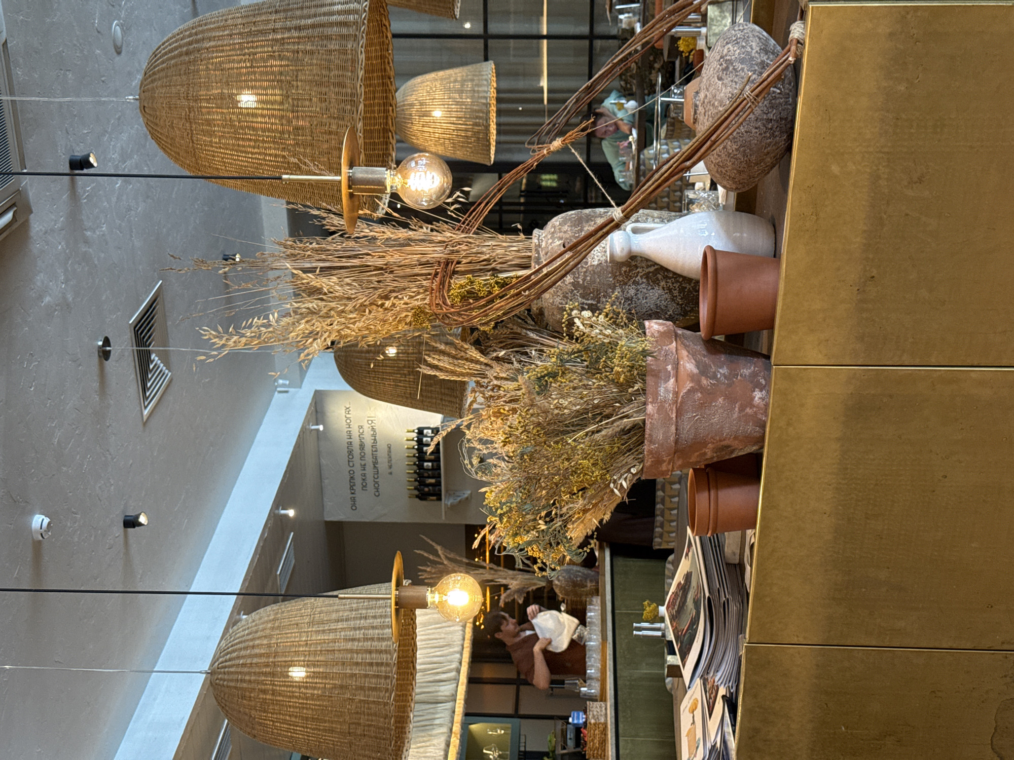 The image shows an interior scene, likely of a restaurant or café in Krasnoyarsk, Russia. In the foreground are several decorative arrangements featuring dried plants and pottery placed on a shelf or counter, which is positioned against a gold-colored panel. Woven light fixtures hang from the ceiling. The interior features large glass panels that give views into the establishment, showing a man with a white cap, and staff wearing aprons, reflected in the glass. A wine rack and a Russian quote can be seen. The scene suggests a stylish and possibly upscale dining establishment with a rustic aesthetic. Magazines and possibly printed menus are partially visible.