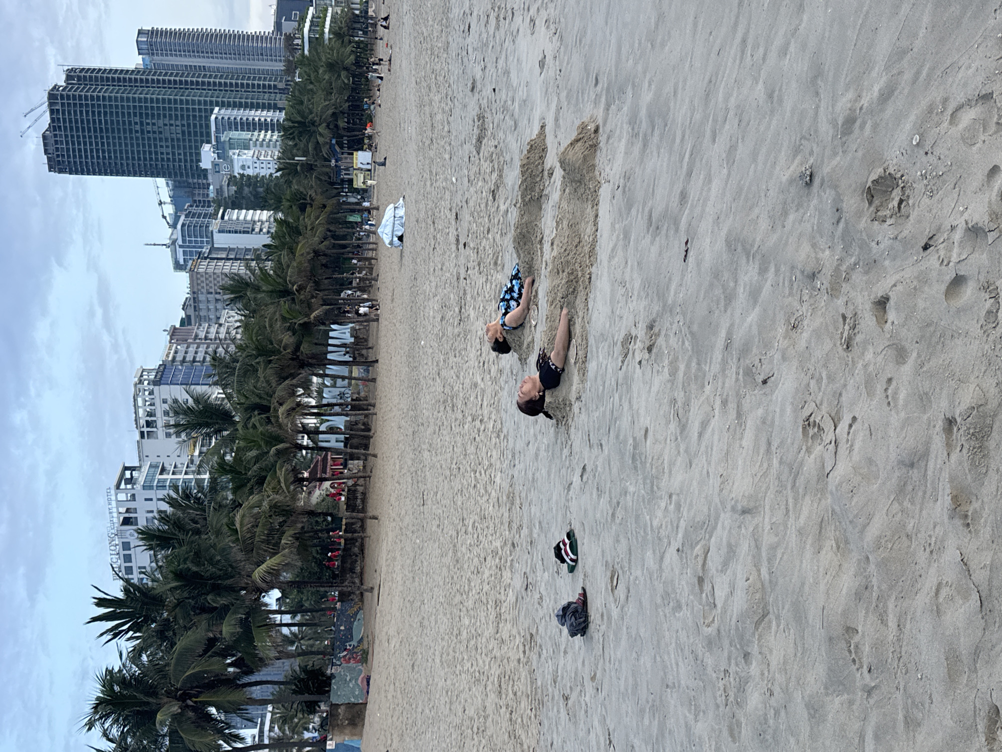 Two people are partially buried in the sand on a beach. Footwear is adjacent to one person. The beach is wide and sandy. A line of palm trees separates the beach from a row of buildings of varying heights, including high-rise buildings. The sky is overcast. The scene is likely in Đà Nẵng, Vietnam.