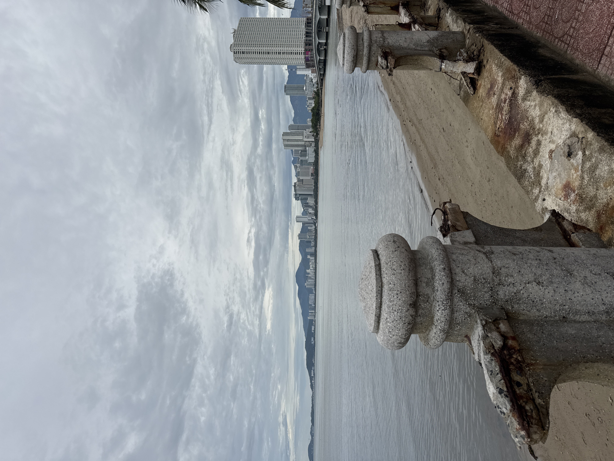 The image depicts a coastal scene in Bac Nha Trang Ward, Vietnam, under an overcast sky. The foreground features a weathered concrete balustrade, showing signs of age and deterioration. Beyond the balustrade, a body of water stretches towards the city skyline, where several tall buildings are visible in the distance. Mountains form a backdrop to the urban landscape. The overall scene suggests a waterfront promenade with an aging infrastructure. There are no people visible in the image.