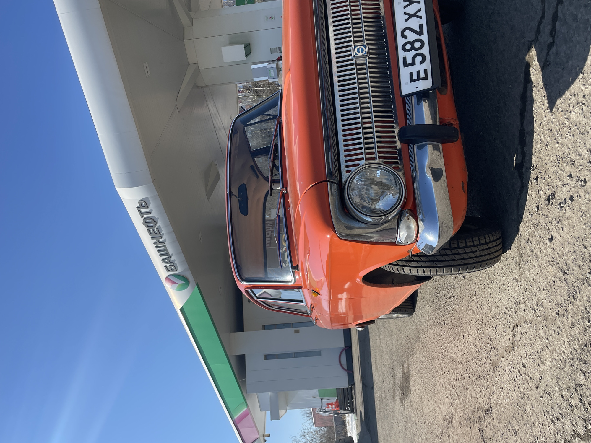 The image depicts a bright orange Moskvich car parked at a Bashneft gas station. The gas station is white with a green stripe and the Bashneft logo is visible on the structure. The license plate number on the car is E582XY. The location is in Izhevsk, Russia. The sky is blue and it appears to be a sunny day. The ground is paved with asphalt. There are no people visible in the frame.