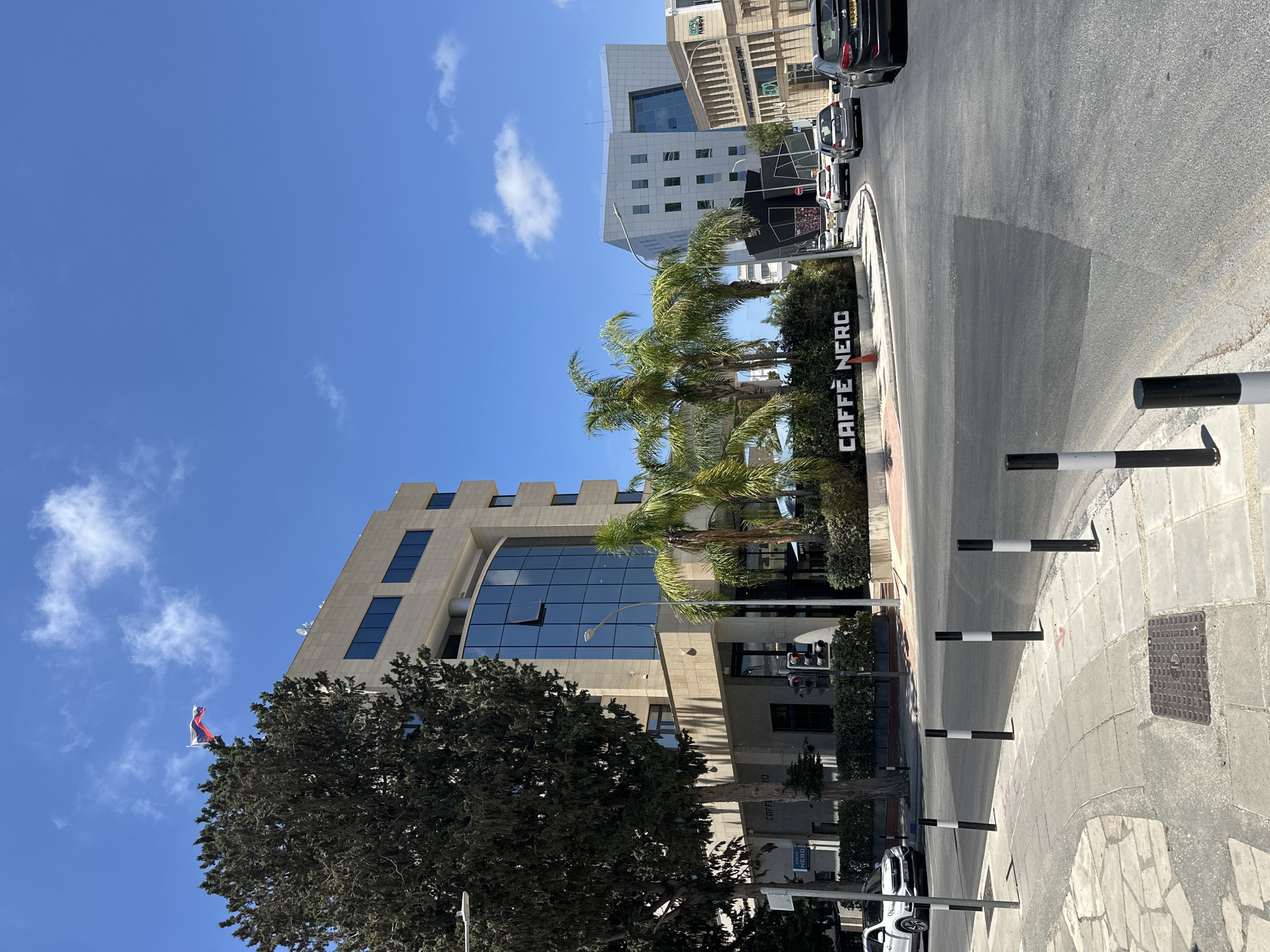 The image depicts a street view in Paphos Municipality, Cyprus, under a clear blue sky with scattered clouds. In the foreground, a paved sidewalk with bollards lines a road where several cars are driving. A white Honda CR-V is parked on the left side of the road.
Dominating the mid-ground is a modern, multi-story building with a beige stone facade and large, dark-tinted windows. A prominent "CAFFE NERO" sign is visible at the base of this building, alongside a lush green hedge and several palm trees. Traffic lights are present at an intersection.
Further in the background, a distinctive, futuristic-looking building with a white, angular facade and glass windows stands out. To its right, another building with a light-colored facade and arched windows is visible. A Russian flag is seen flying from a flagpole atop the beige stone building. The scene conveys a sense of urban activity and modern architecture.