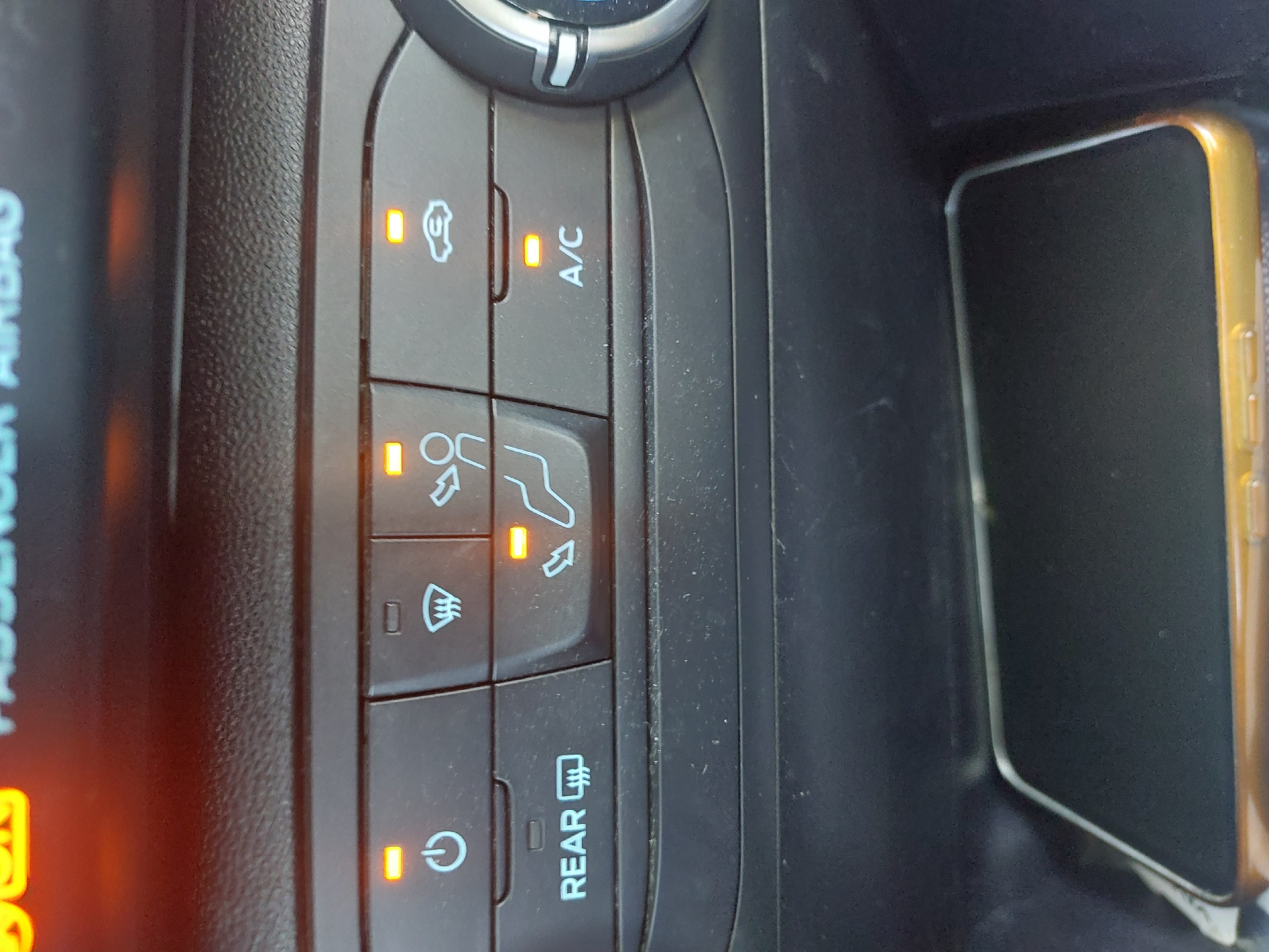 The image depicts a car's climate control panel. The panel includes buttons for recirculation, A/C, seat heating, windshield defrost, and rear defrost. Indicator lights are illuminated for recirculation, seat heating, windshield defrost, and power. A mobile phone lies in the car's center console. The location of Ravadinovo, Bulgaria, is not discernable from the image.
