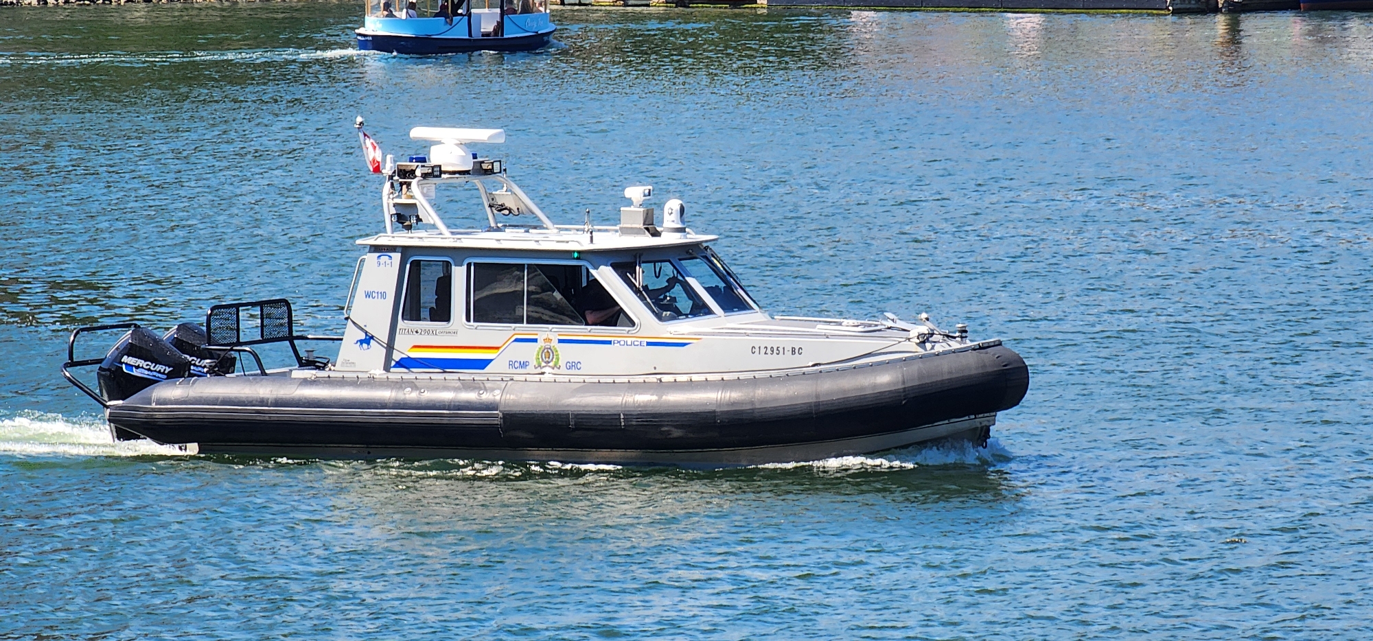 An RCMP (Royal Canadian Mounted Police) rigid-hulled inflatable boat (RHIB) with two Mercury outboard motors is cruising on a body of water under a clear sky. The boat, identified by the marking "WC110" and "C12951-BC", features a white cabin with tinted windows and police markings, including the RCMP crest and the word "POLICE". A Canadian flag is mounted on a pole at the top of the cabin, along with various communication and surveillance equipment. The boat is generating a wake, indicating movement. In the background, another smaller blue boat with passengers is visible, as is a distant shoreline with buildings. The water is choppy and blue, reflecting the bright sunlight. The overall scene depicts a police patrol on the water.