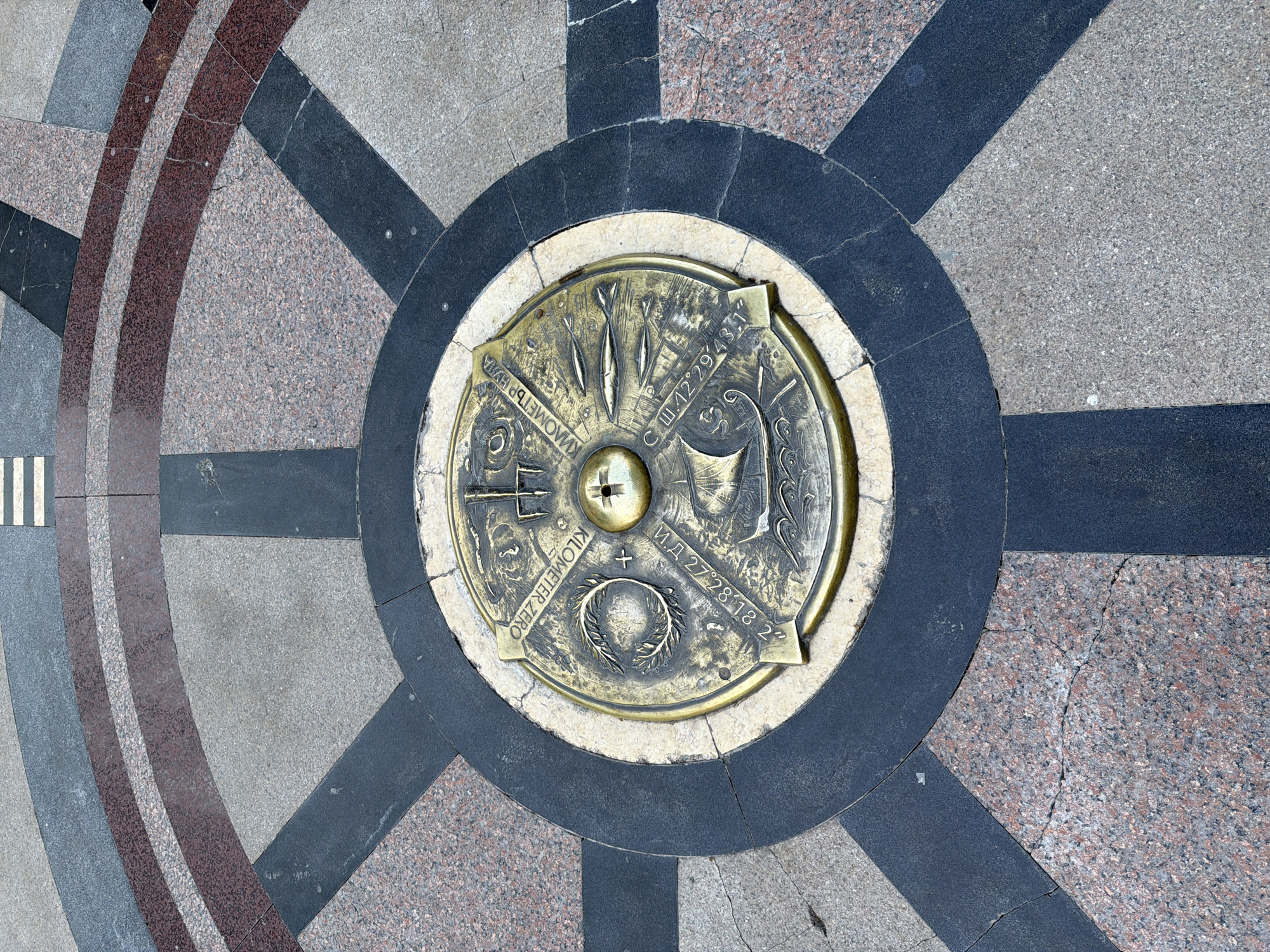 This is a detailed, overhead shot of a brass "Kilometer Zero" plaque set into a circular stone mosaic on a ground surface. The mosaic consists of alternating dark gray and light gray trapezoidal stone pieces, with a partial border of red stone on the left side. The plaque itself has a central, slightly raised hub with a small cross design. Various engraved symbols and text are visible around the central hub. The text is written in Cyrillic and Roman alphabets. The inscription "КИЛОМЕТЪР НУЛА" and "KILOMETER ZERO" can be read. Other engravings include a group of three fish-like figures and the coordinates "С Ш.42°29′43,1" and "И.Д 27°28′18 2"". A small wreath is visible at the bottom of the plaque. The ground surface appears to be an outdoor public space, and the mosaic seems to be part of a larger design. The scene is located in an urban environment, although the specific city and location are unknown.