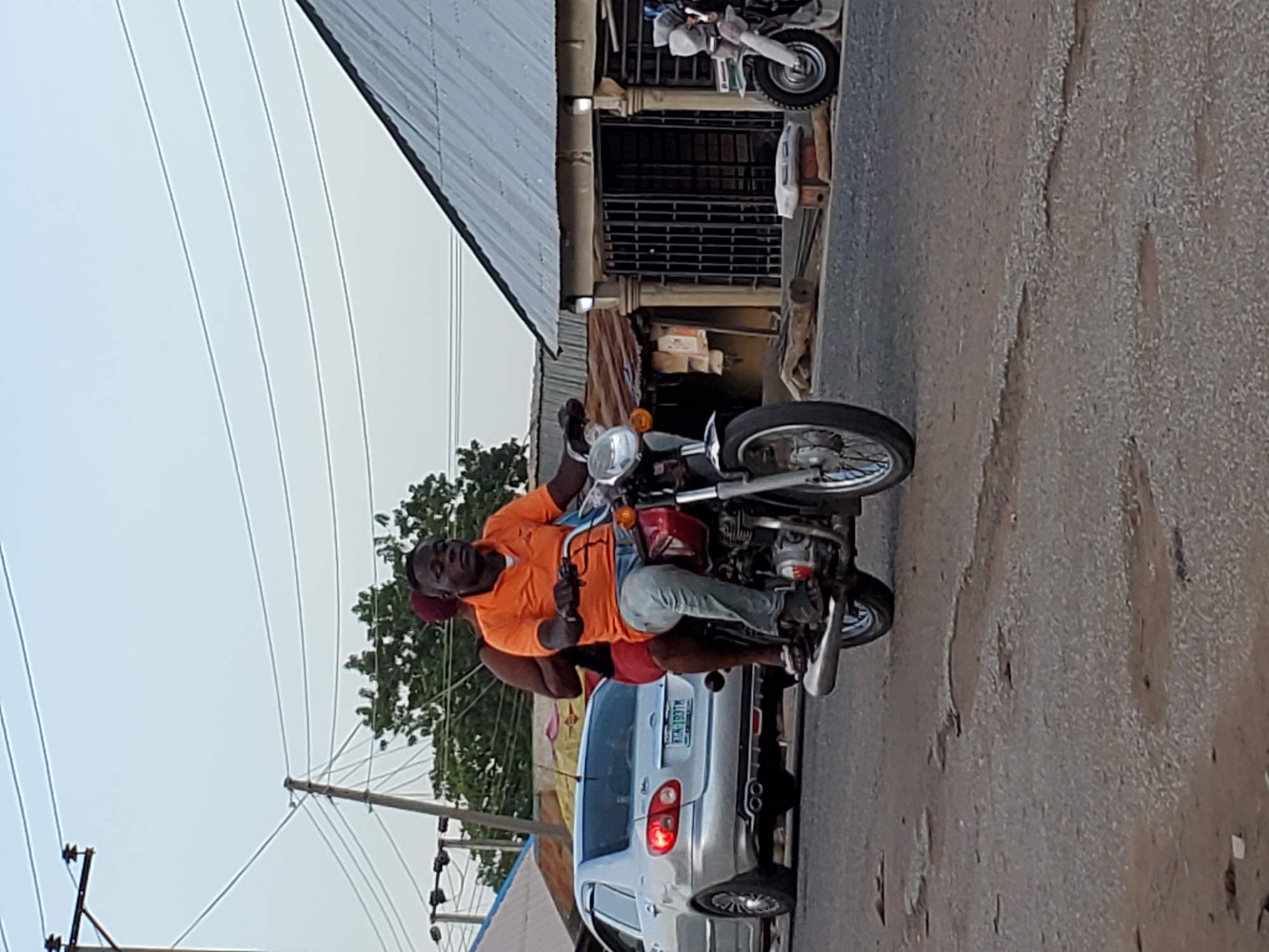 This portrait-oriented image captures a street scene in Mararaba, Nigeria, featuring a man on a motorcycle. The main subject is an adult male, wearing an orange short-sleeved polo shirt and light-colored jeans, operating a red and chrome motorcycle. A second individual, largely obscured and wearing a reddish garment, is seated behind him on the motorcycle. The driver has a neutral expression and is looking forward.
Immediately behind the motorcycle, a silver four-door sedan is visible, either parked or moving slowly. The car's rear license plate, "KJA-187TM," is clearly legible. The road surface appears to be unpaved or heavily worn asphalt, showing signs of unevenness and use.
In the background, buildings with corrugated metal roofs can be seen, suggesting a commercial or residential area. Overhead, multiple power lines stretch across a bright, clear sky, indicating daytime. Green foliage from trees is visible, adding to the natural elements of the setting. The scene depicts a common mode of transport and daily life in a bustling urban or semi-urban environment.