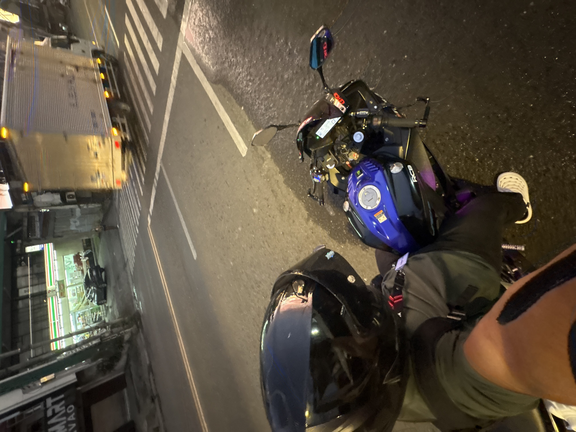A first-person perspective shot shows a rider seated on a motorcycle at a street intersection in Davao City, Philippines. The rider is wearing a black helmet, a gray shirt, and black pants with white shoes. The motorcycle has a blue and black fuel tank. In the background, a 7-Eleven convenience store is visible with a black car parked outside. A large white truck is crossing the intersection in the distance. The street is wet, indicating recent rain, and streetlights illuminate the scene.