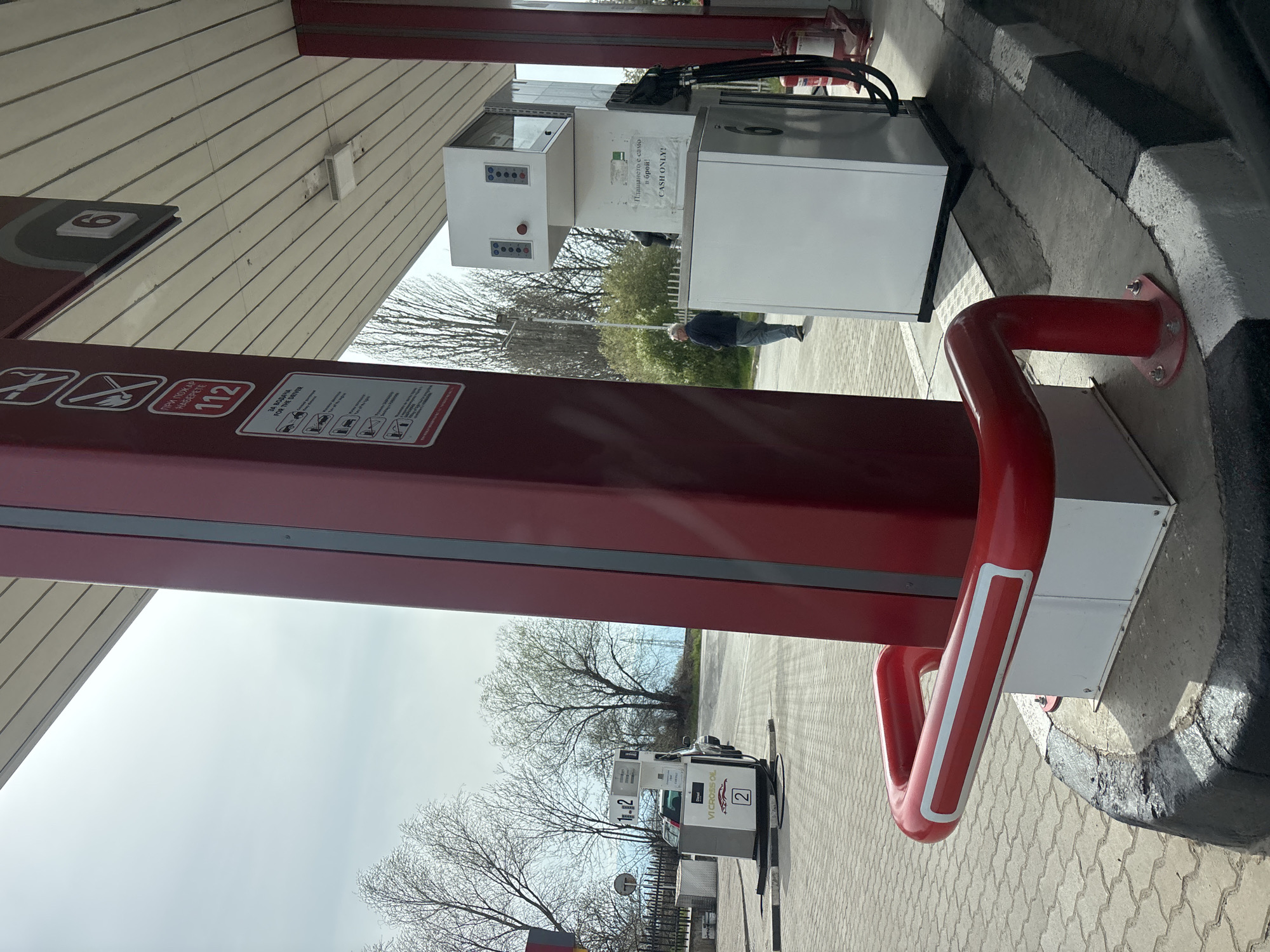 The image shows the interior of a gas station canopy in Dolni Bogrov, Bulgaria. One can spot red support pillars, signage displaying safety instructions, and gas pumps. One pump is labeled "VI CROSS OL," and another displays the message "CASH ONLY!". An individual is walking in the background. The surrounding environment includes paved surfaces, trees, and a cloudy sky.