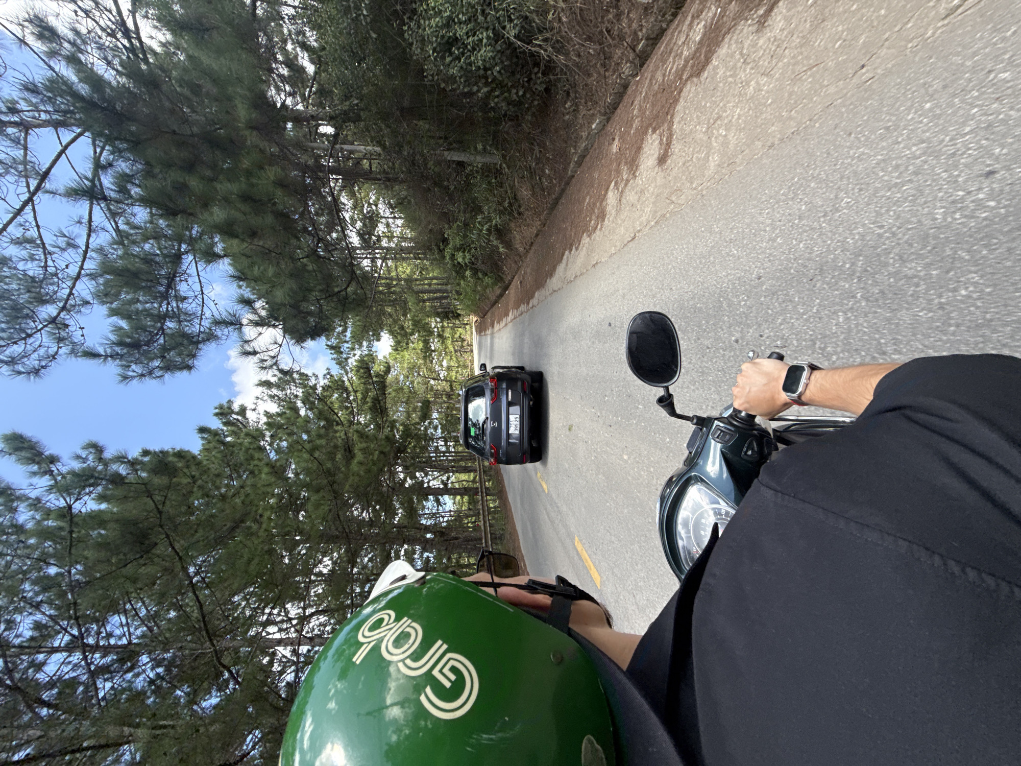 The image shows a first-person perspective from a scooter on a paved road lined with pine trees, likely in the city of Lạc Dương, Vietnam. The road is asphalt with a yellow line marking the lane. In the distance, a dark-colored SUV is driving ahead on the same road. The sky is blue with scattered white clouds. The foreground shows the rider's green helmet with the "Grab" logo, a black shirt, and a smartwatch on the right wrist. The scooter's handlebars and dashboard are partially visible. The surrounding environment is densely populated with tall pine trees, and there is a concrete drainage ditch along the side of the road.
