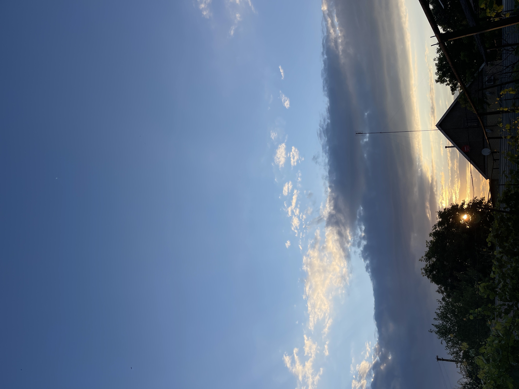 The image captures a sunset scene in the city of Sredny, Russia. The sky is a blend of blue and light orange hues, with scattered clouds. In the foreground, a house with a pointed roof and an antenna is visible, partially silhouetted against the bright sky. Lush green trees and foliage surround the house. The setting sun casts a warm glow, peeking through the branches of a large tree on the right side of the frame. The overall impression is one of a tranquil evening in a rural or suburban setting.