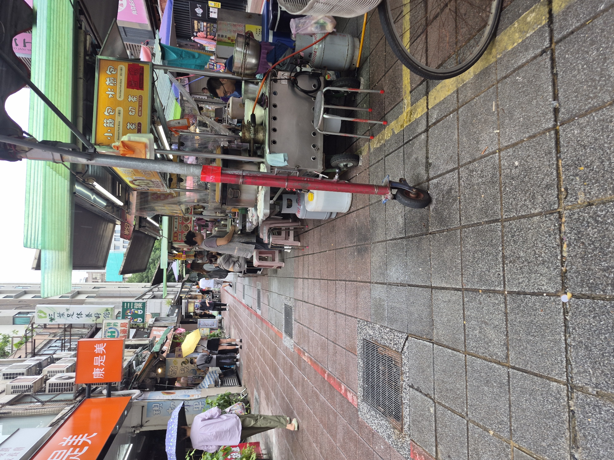 This is a street-level, eye-level shot of a bustling market or shopping street in Taipei, Taiwan. The scene is filled with various food stalls and shops lining both sides of a pedestrian walkway. In the foreground, a red and blue wheeled scooter is parked on the grey tiled ground, which is marked with a yellow line. To the right, the rear wheel of a bicycle is visible.
Several people are present in the scene. On the left, a person in a light purple shirt and green pants walks away from the camera, holding a purple umbrella. Further down the street, other pedestrians can be seen browsing stalls or walking. On the right, in front of a food stall with a yellow sign featuring Chinese characters and English text like "Steamed Pork Buns" and "Milk Tea," a man in glasses is working behind the counter, attending to customers. He appears to be serving food. The stalls are covered by green awnings and lit by fluorescent lights. Behind the stalls, multi-story buildings with air conditioning units are visible. The ground is paved with square grey tiles, interspersed with occasional metal grates. The overall impression is a lively urban environment with a focus on street food and commerce.