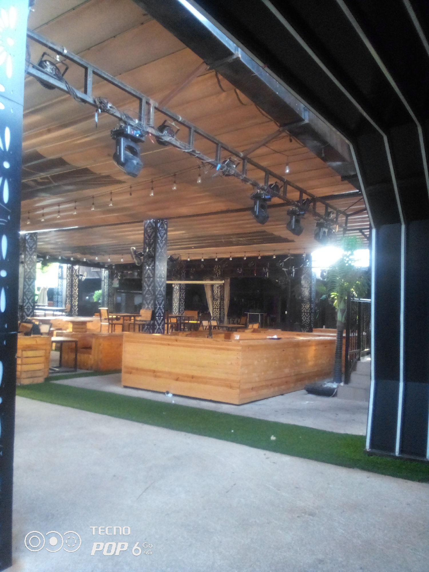 The image shows an outdoor seating area that appears to be part of a restaurant or entertainment venue in Howayi, Nigeria. The area is covered by a wooden ceiling structure, which is supported by pillars. Several stage lights are suspended from a metal truss system above the seating. The seating itself consists of low wooden benches and tables arranged on what appears to be artificial turf. In the background, more seating and architectural elements are visible, including decorative pillars and what looks like a stage or performance area. A potted plant is visible to the right. The overall impression is of a well-lit, modern outdoor venue.