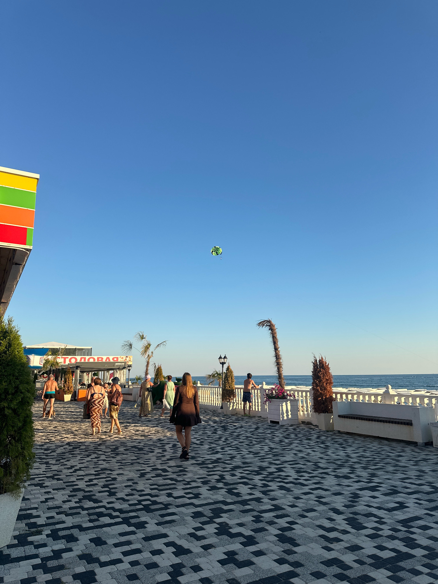 A bright, sunny day in Lermontovo, Russia, unfolds on a paved promenade leading to the Black Sea. In the foreground, a woman in a dark dress walks away from the camera. The promenade is laid with a distinctive pattern of black and grey interlocking pavers. To the left, a building with vibrant orange, yellow, and green stripes is partially visible. Further down the promenade, several people are seen strolling, some in swimwear. A "СТОЛОВАЯ" sign, indicating a canteen, is visible above a structure. Palm trees and decorative bushes dot the landscape. In the distance, the calm blue sea meets the horizon under a clear blue sky. A green and yellow parasail floats high in the sky, a testament to recreational activities offered in this seaside town. A white railing with decorative balusters runs along the edge of the promenade, separating it from a series of white loungers, suggesting a beach resort area.