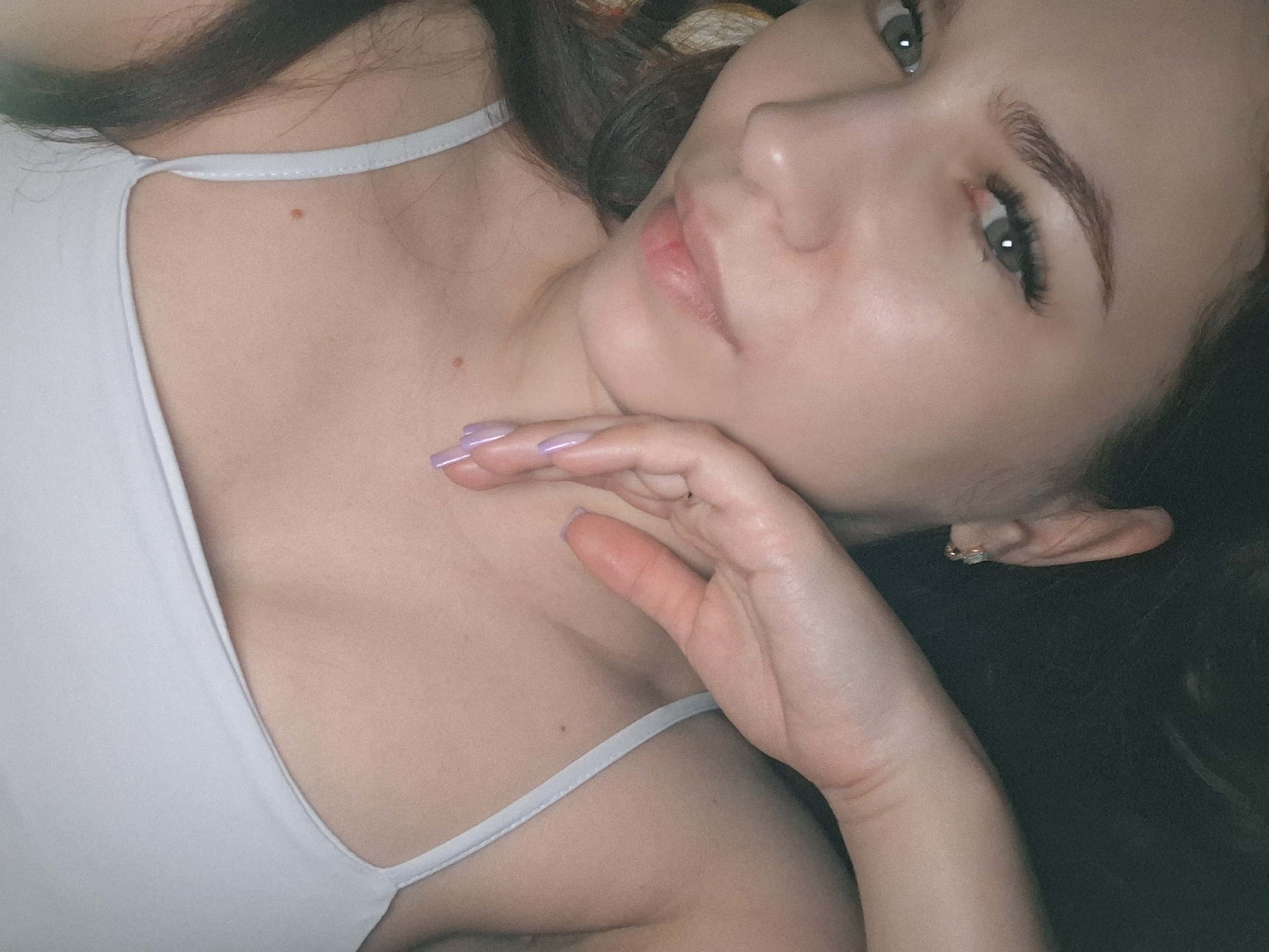 A close-up portrait of a woman with fair skin and brown hair. She is wearing a light-colored tank top with thin straps. Her hand is near her face. She has an earring in her left ear. Her fingernails are painted a light purple color. The location is not discernible from the image.
