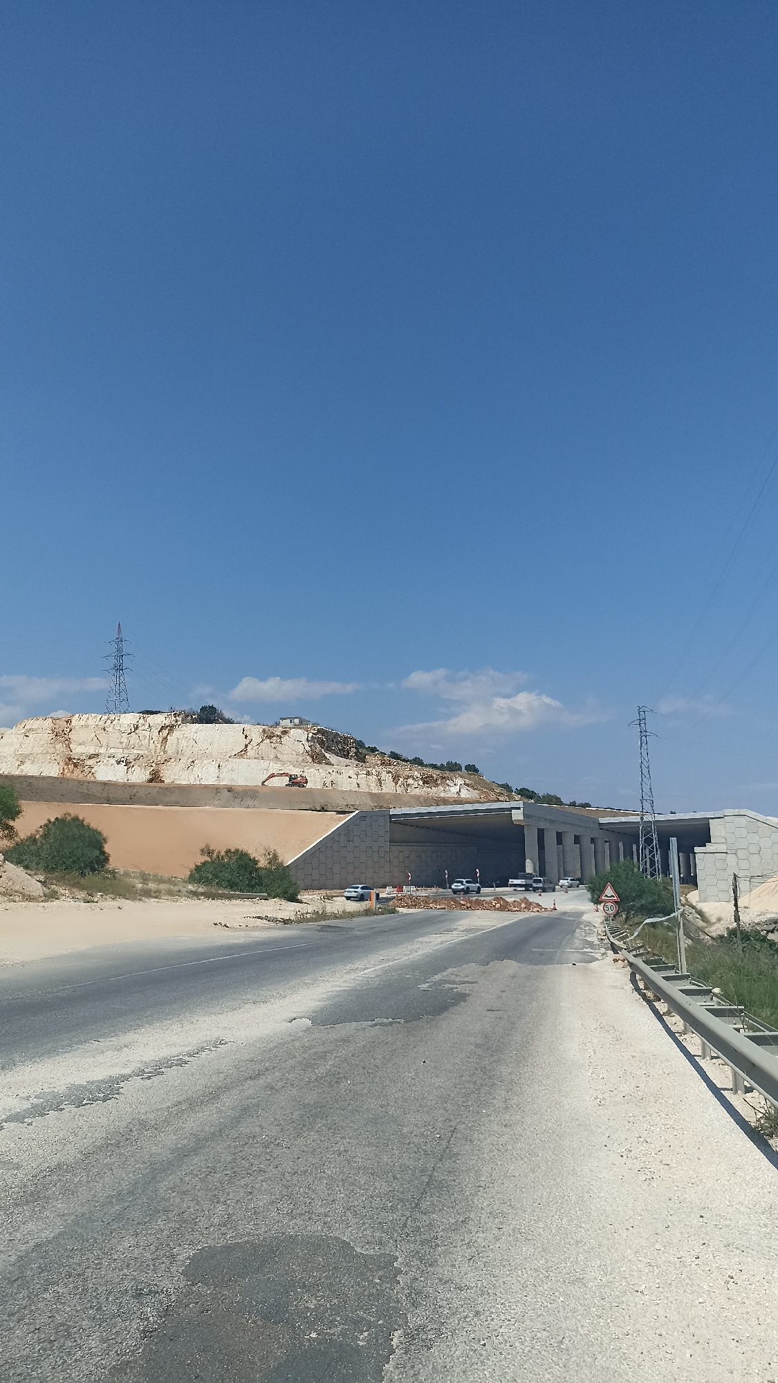 The image depicts a road leading to a partially constructed tunnel. The road surface is worn with patches of discoloration and damage. A concrete barrier runs along the right side of the road. To the left, there is a sandy area with sparse vegetation and a light-colored, rocky hillside. An excavator is visible on the hillside. The tunnel structure is made of concrete and has multiple openings. Several vehicles are visible either entering or near the tunnel. A speed limit sign displaying "50" is on the right side of the road near the tunnel entrance. Overhead power lines and towers are present in the background. The sky is blue with scattered clouds. The location is Unknown, Unknown.