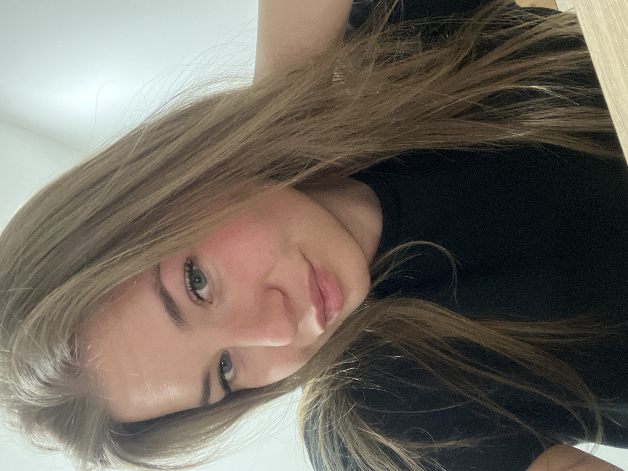 A close-up, selfie-style photograph shows a woman with light brown, long hair, wearing a black shirt. Her face is tilted towards the camera from an angle. She has fair skin, grey eyes, and light pink lips. There is no indication in the image that it was taken in Budva, Montenegro, or any other specific city. The background is a plain white wall and a light-colored wooden surface.