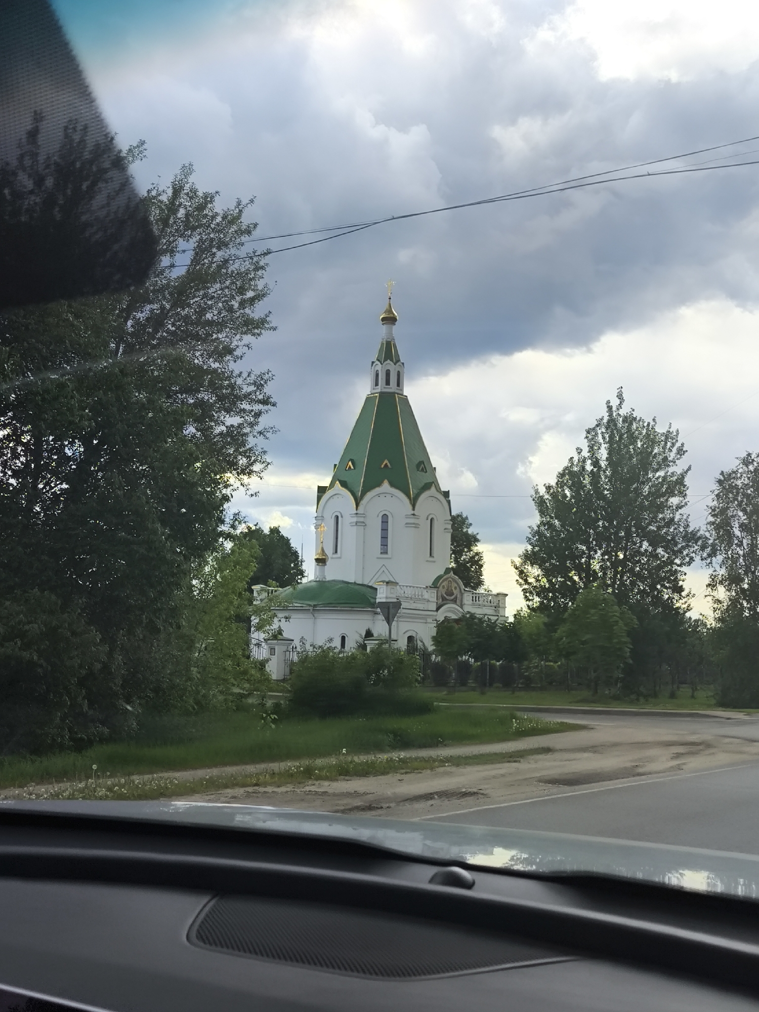 A white building with a green roof and golden domes is visible from inside a car. The building is surrounded by green trees and grass. The sky is cloudy. The location of the image is Unknown, Unknown.